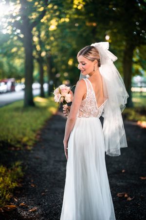 Bride in lace back wedding gown holding bouquet on tree-lined path.