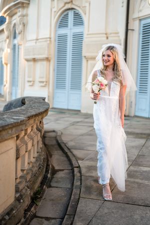 Bride in white lace gown holding bouquet against historic building with blue shutters.