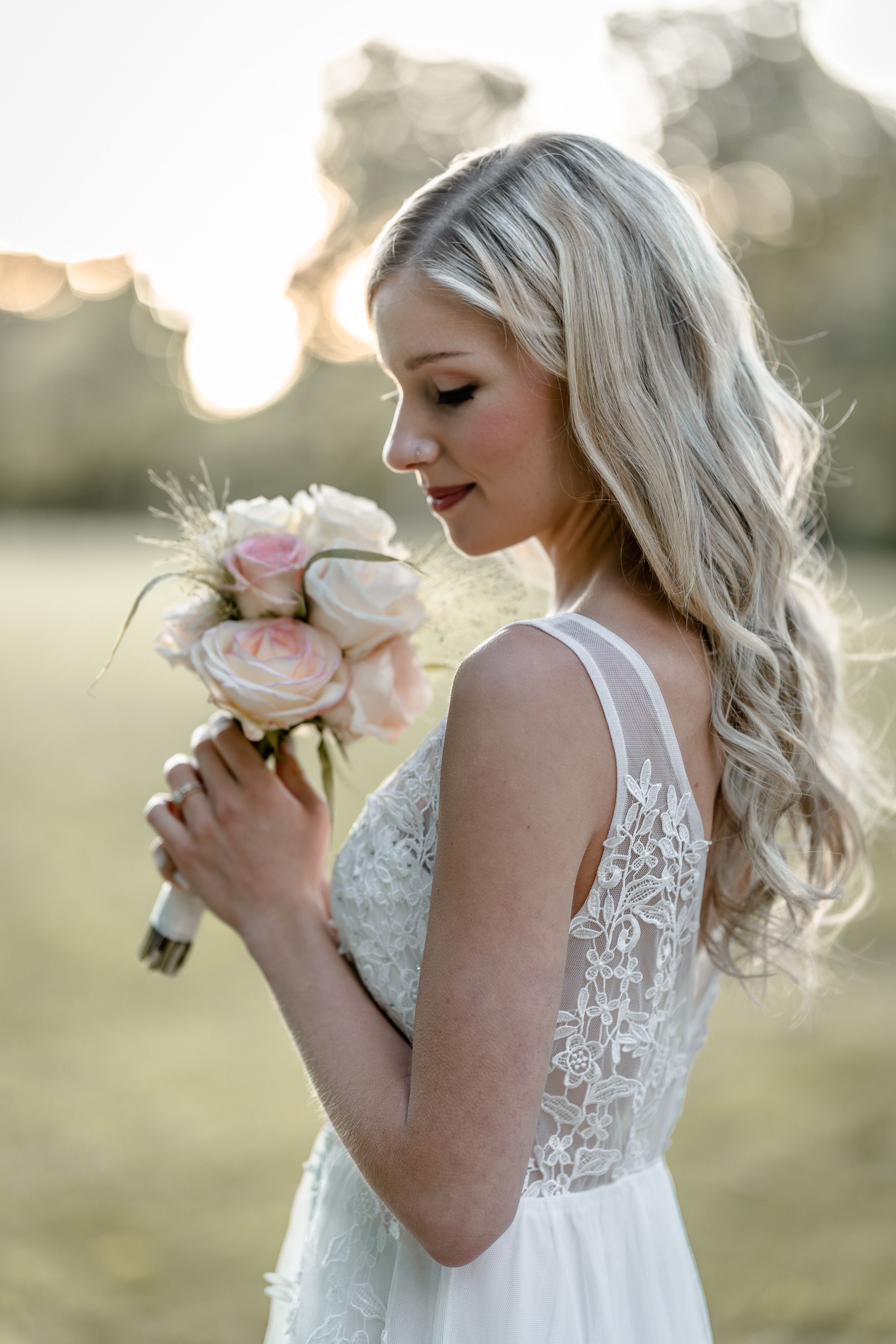 Bride in lace wedding dress holding pink rose bouquet during golden hour portrait session.