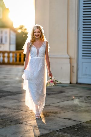 Bride in lace wedding dress walking at sunset holding bouquet.