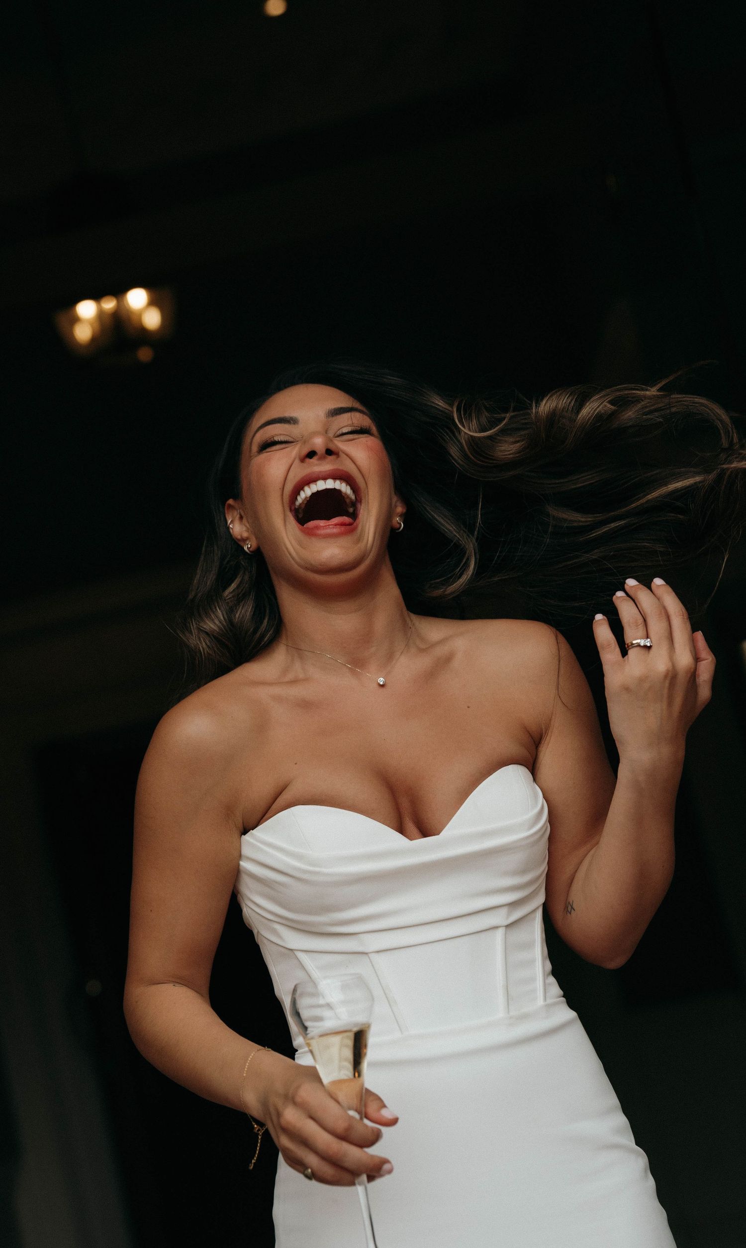 A joyful candid moment captured in white strapless dress with hair flowing and genuine laughter in a dark setting.