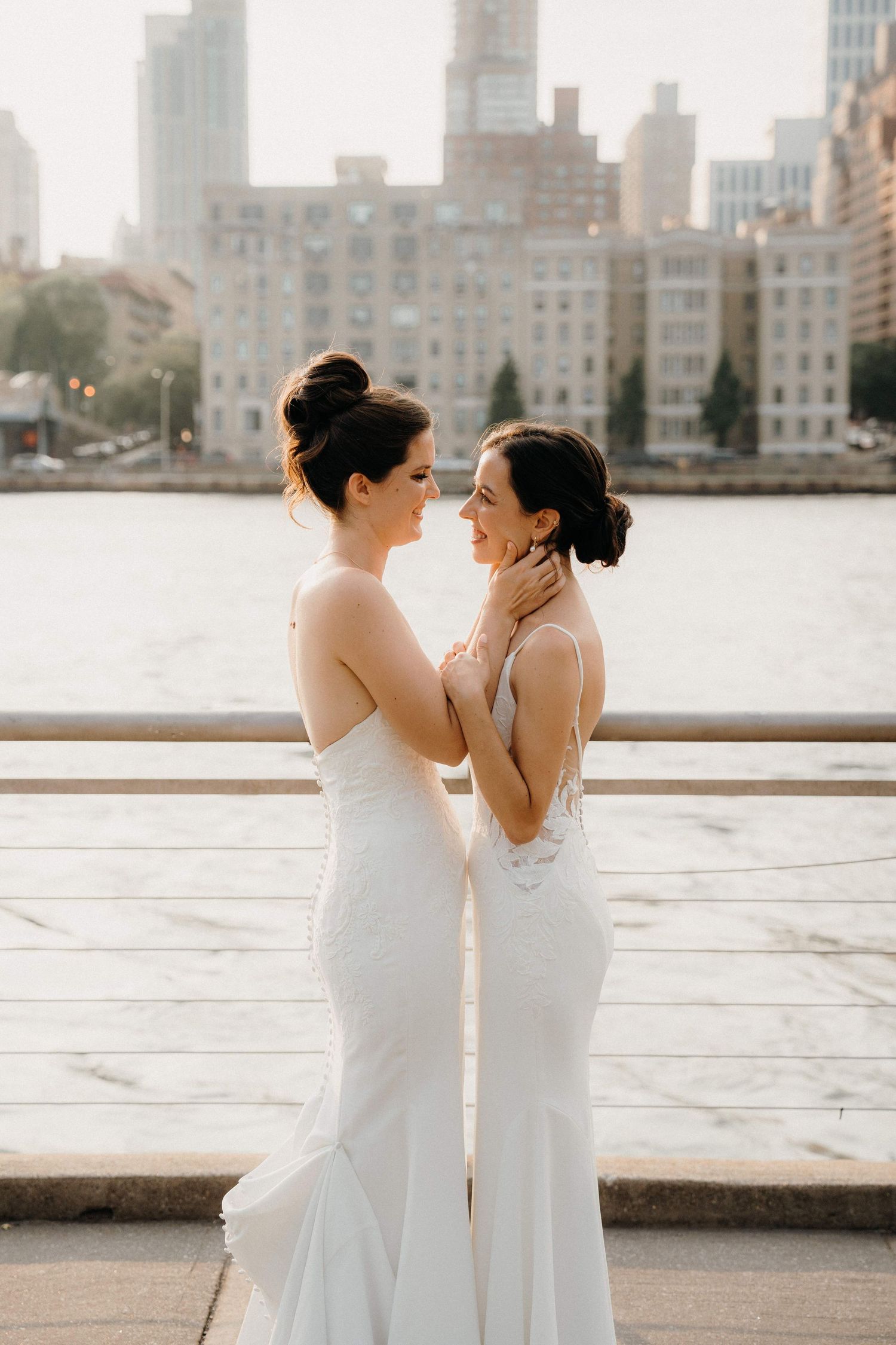 Two women in white dresses share an intimate moment against a city skyline at the Sanctuary Roosevelt Island NYC