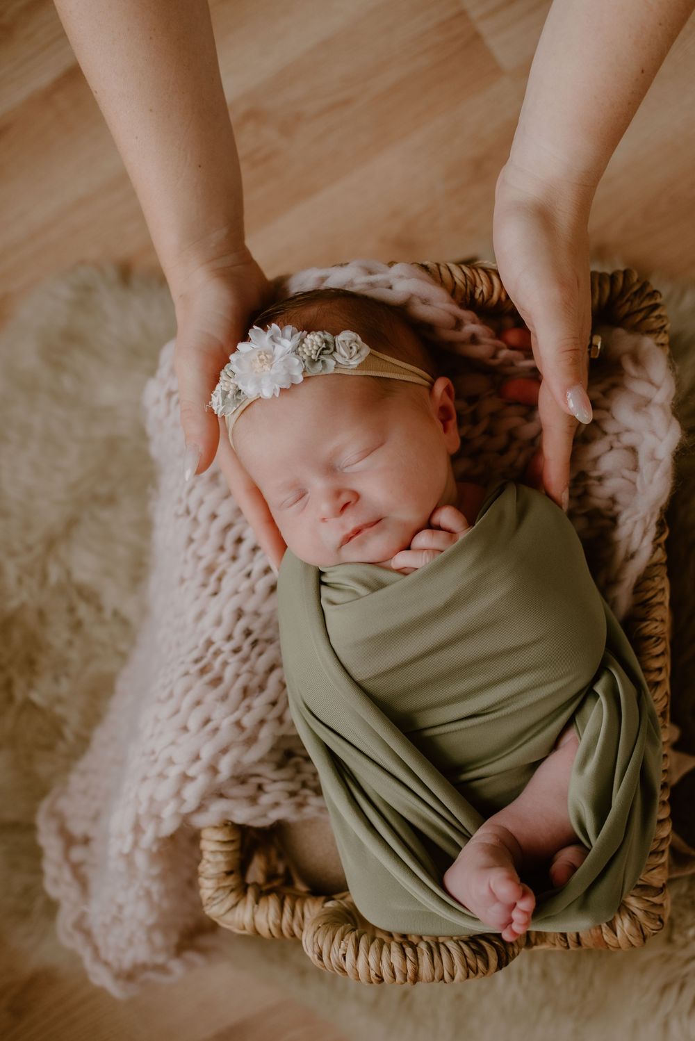 Sleeping newborn wrapped in olive green swaddle with delicate headband in woven basket.