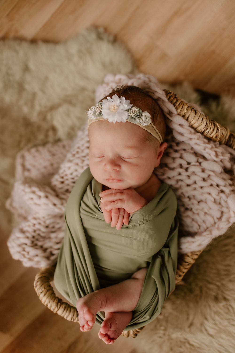 Newborn baby wrapped in sage green fabric on cream knit blanket with floral headband.