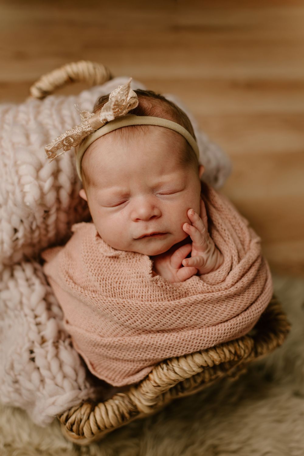Newborn wrapped in pink knit blanket sleeping in woven basket with decorative headband.