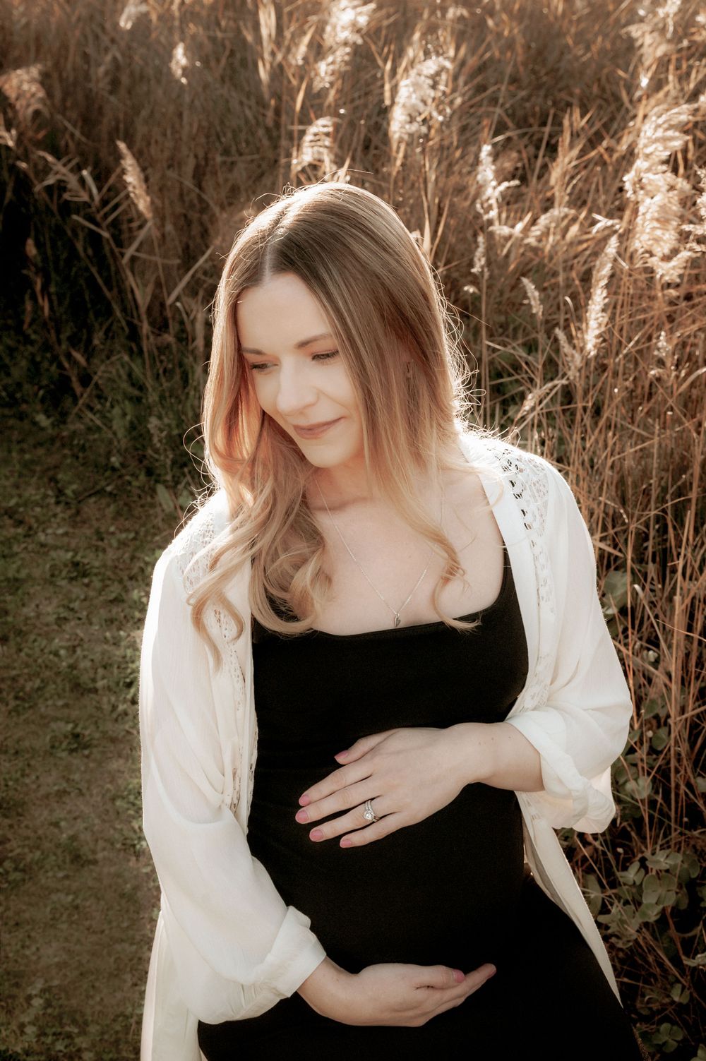 Expectant mother in black dress and white cardigan poses in front of tall grass at sunset.
Altona Family Photographer Melbourne