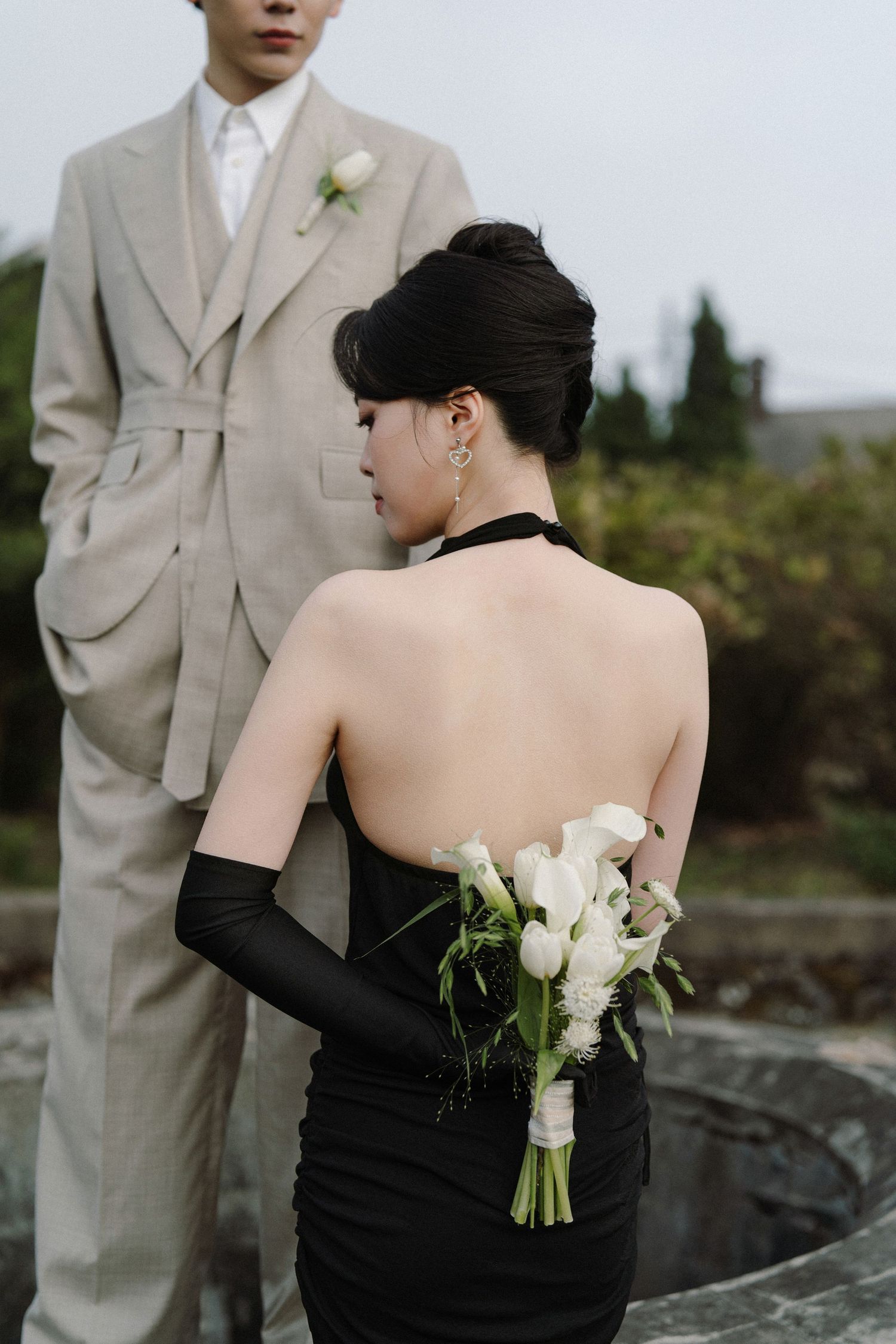 Wedding attire featuring a beige suit and black strapless dress with white roses on rocky terrain.