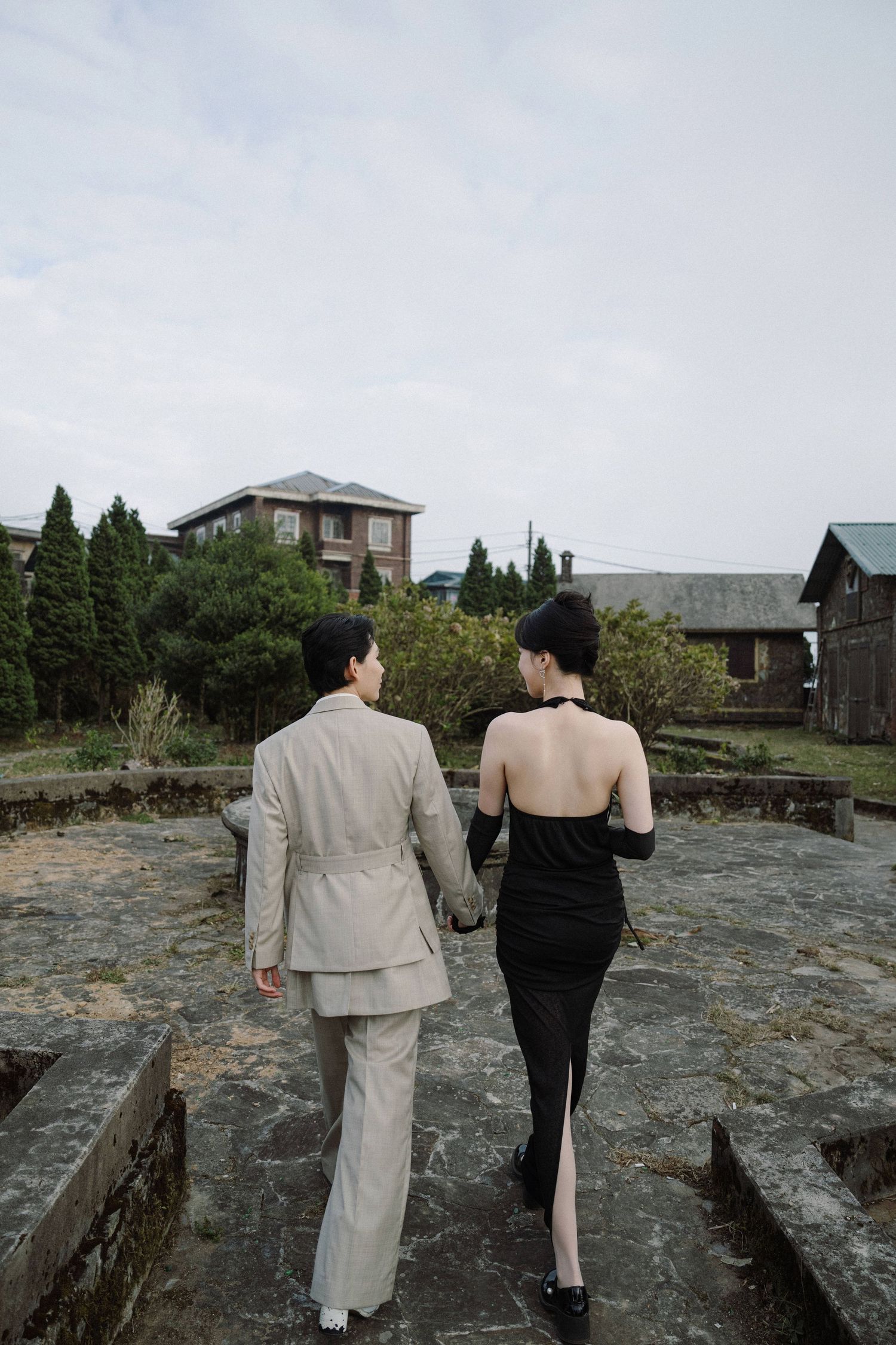 A couple in formal wear walking together on a stone path with buildings in the background.