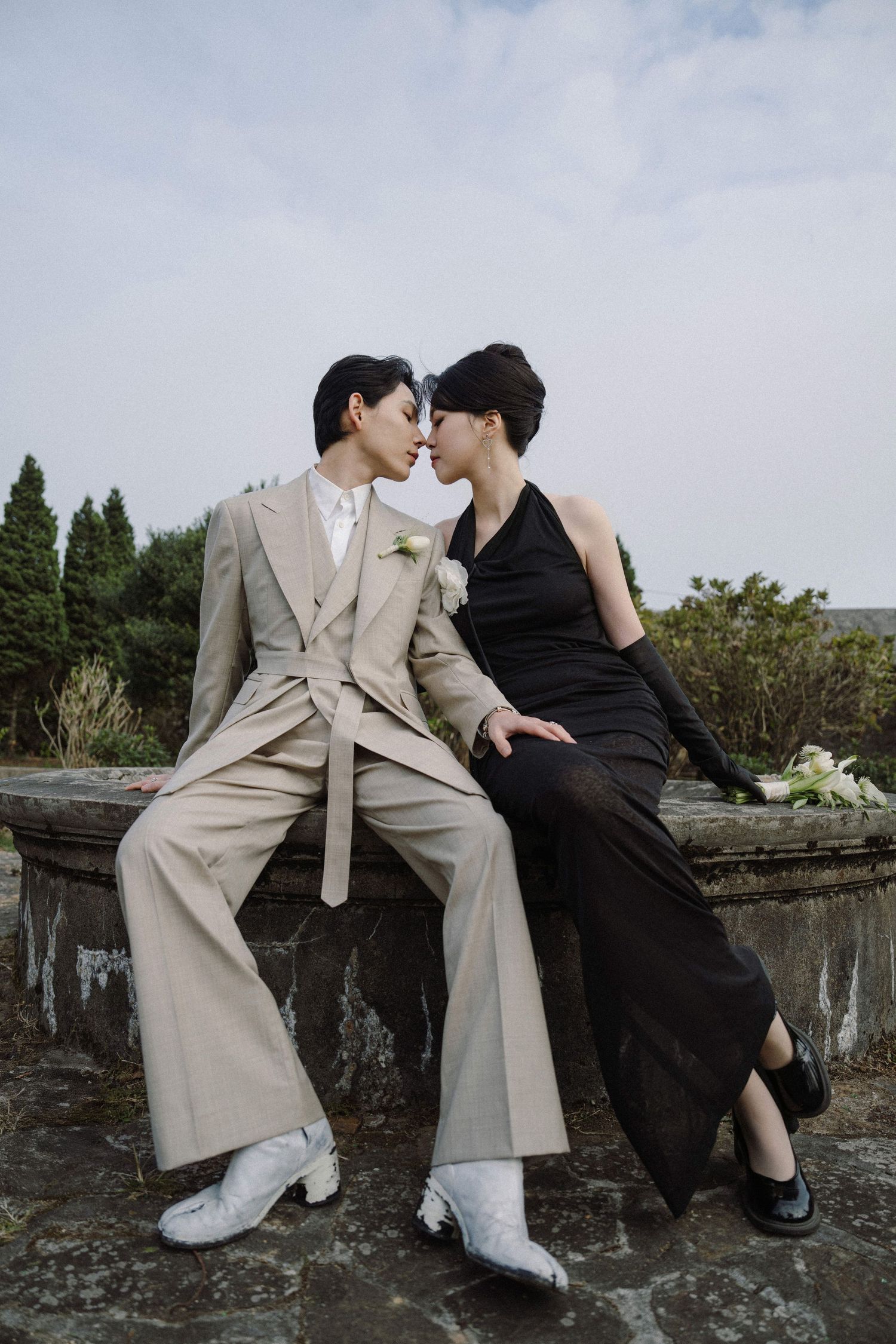 Romantic couple in contrasting beige and black outfits sitting on a stone wall.