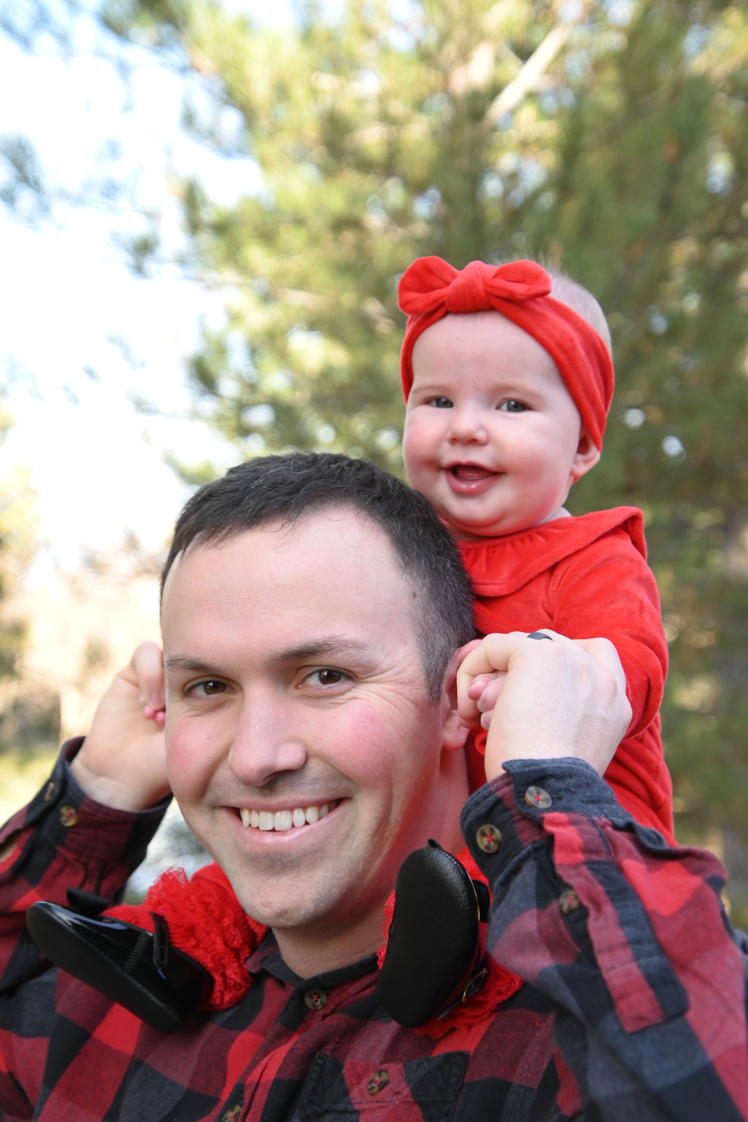 A happy moment outdoors with someone wearing a red buffalo plaid shirt holding a baby in bright red clothing.