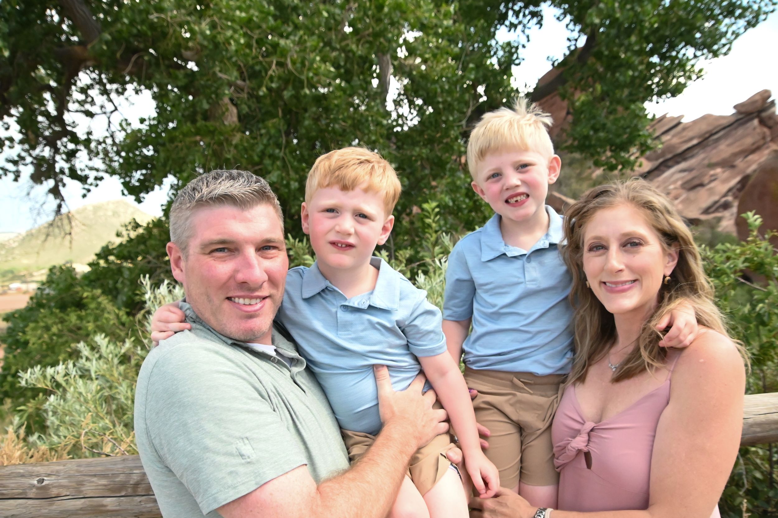 A family of four poses together outdoors with red rocks and green trees visible in the background.