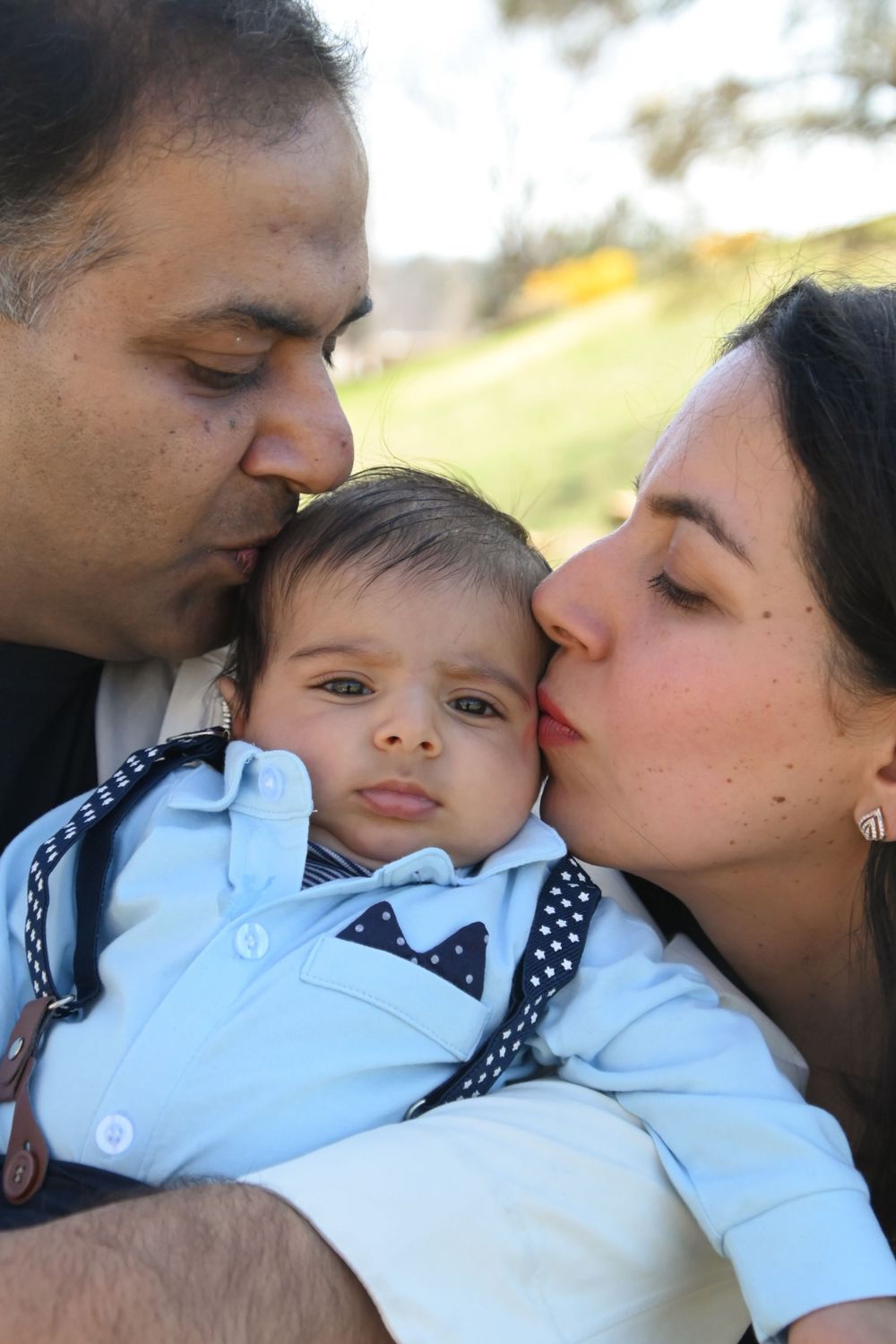 Parents lovingly kiss their baby wearing a light blue formal outfit while outdoors on a sunny day.