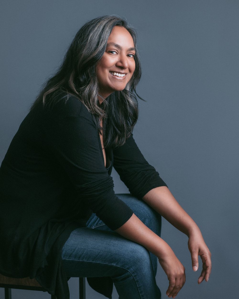 Studio portrait with subject in black top and jeans sitting informally against grey background. Mika Larson Photography.