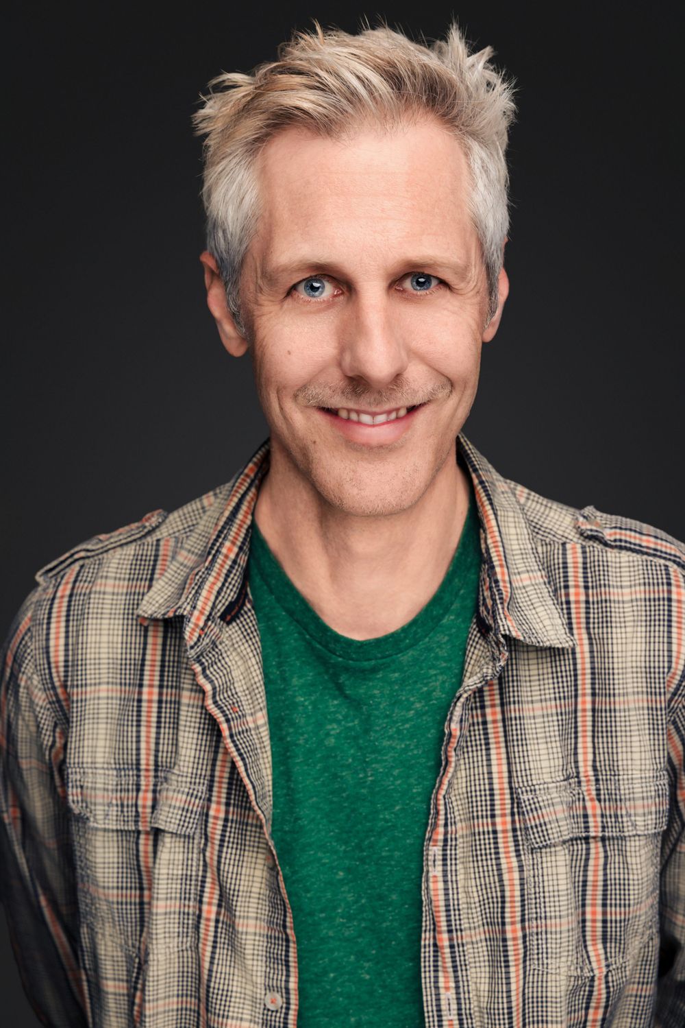 A friendly person wearing a plaid blazer and green t-shirt smiles against a dark background with styled gray hair.