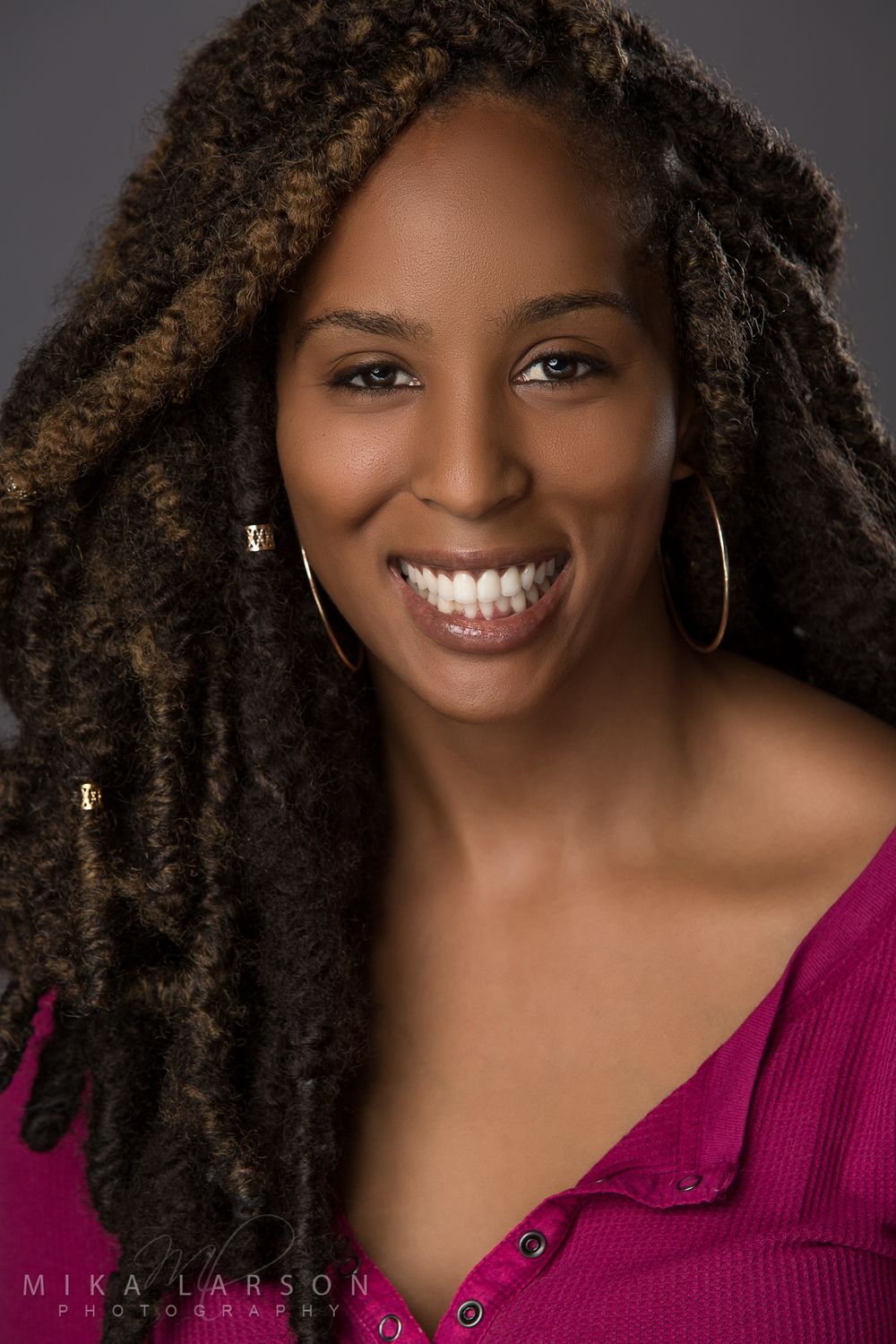 Actor headshot of person with long locs hairstyle wearing magenta top and hoop earrings. Mika Larson Photography.