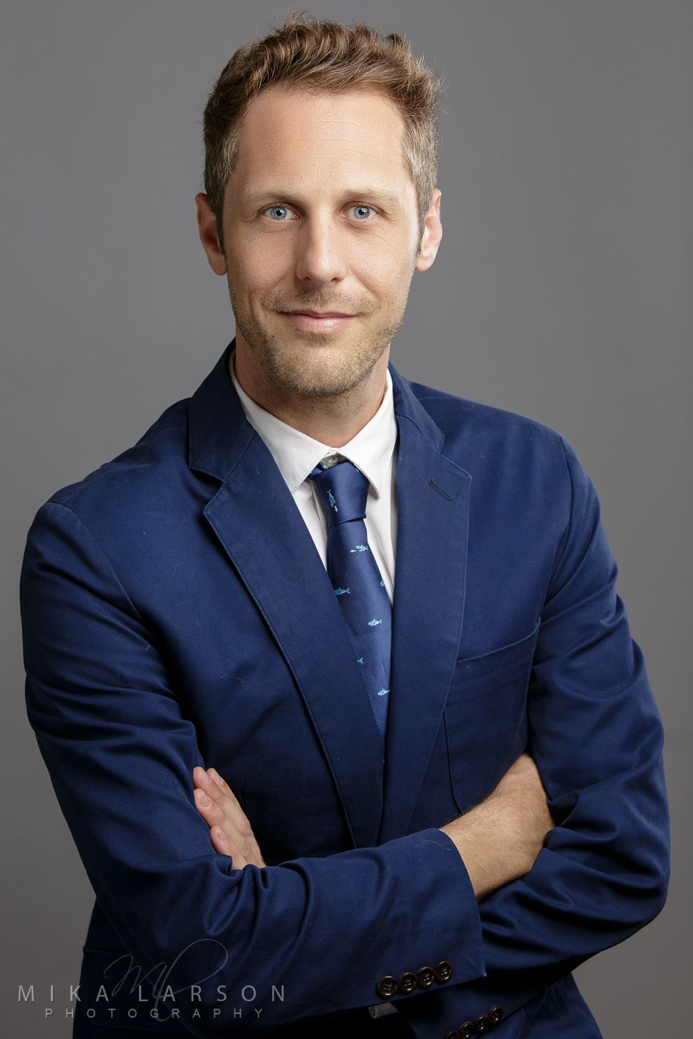 Corporate headshot of person in navy suit and tie against gray background. Mika Larson Photography.