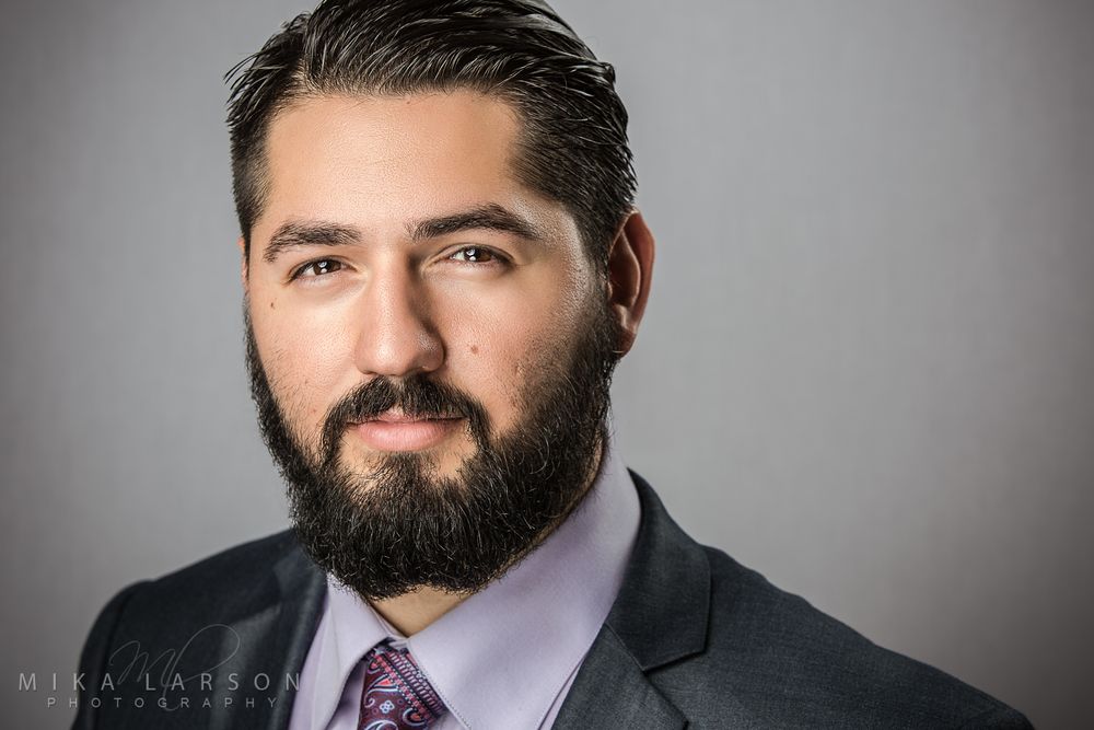 Professional headshot of person with beard wearing suit and purple tie against gray background. Mika Larson Photography.