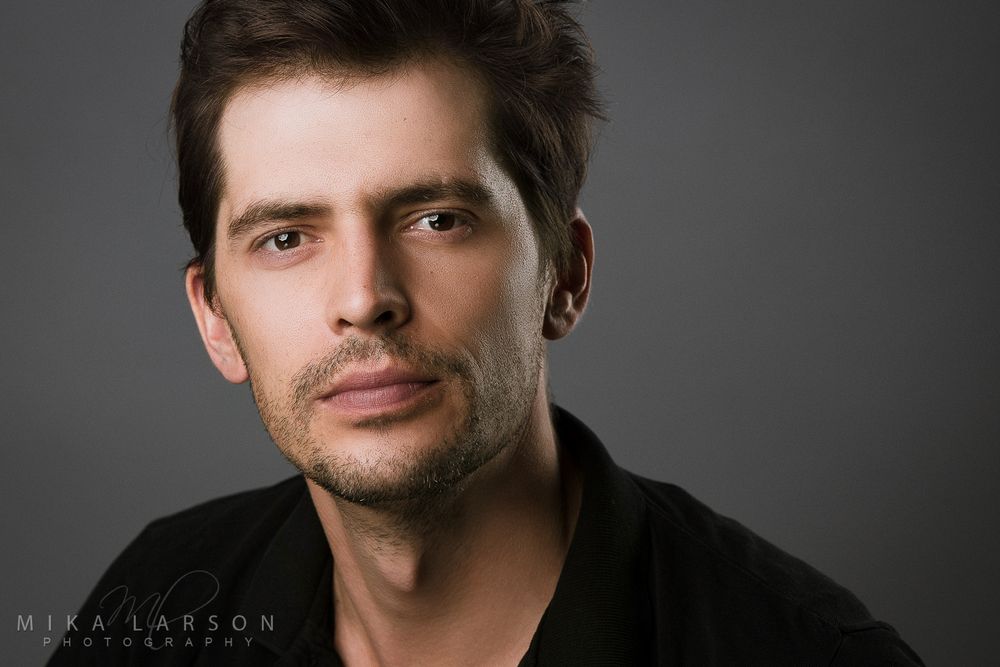 Studio headshot of person with stubble wearing black shirt against dark background. Mika Larson Photography.