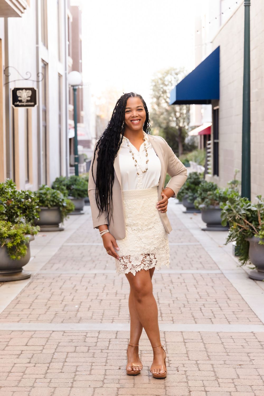 A person wearing a white lace dress and beige cardigan stands smiling on a brick-paved street lined with planters.