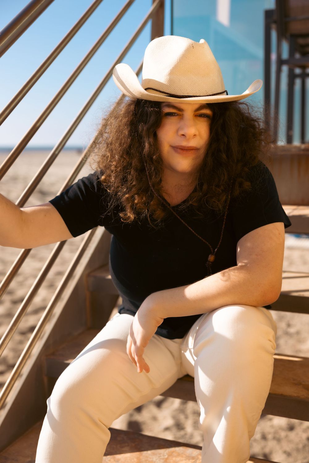 Someone with long curly dark hair wearing a black t-shirt, white cowboy hat, and light pants sits on outdoor stairs.