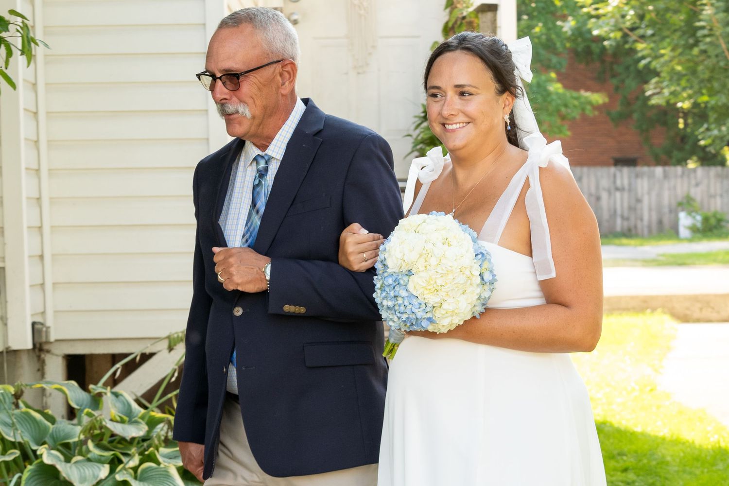Two elegantly dressed people embrace during an outdoor wedding ceremony in front of a white house.