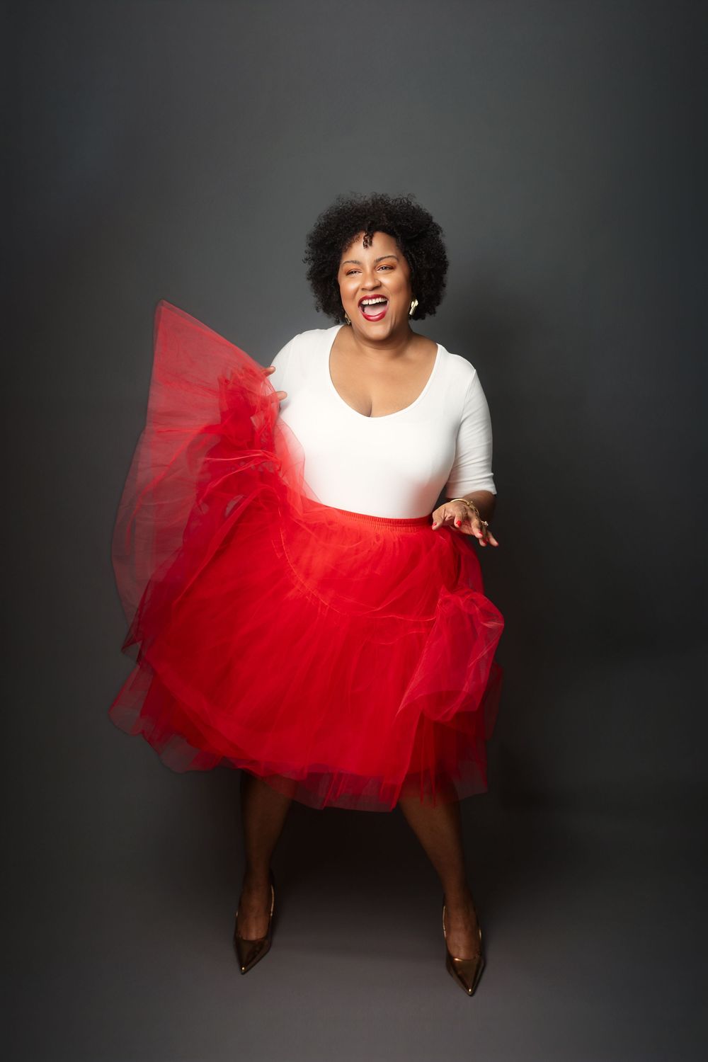 Someone wearing a crisp white top and voluminous red tulle skirt poses expressively against a dark backdrop in a photo series.