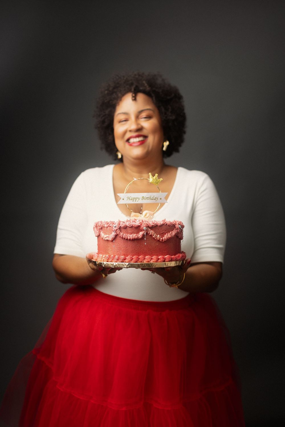 Person wearing white top and red tulle skirt holds pink birthday cake against dark background.