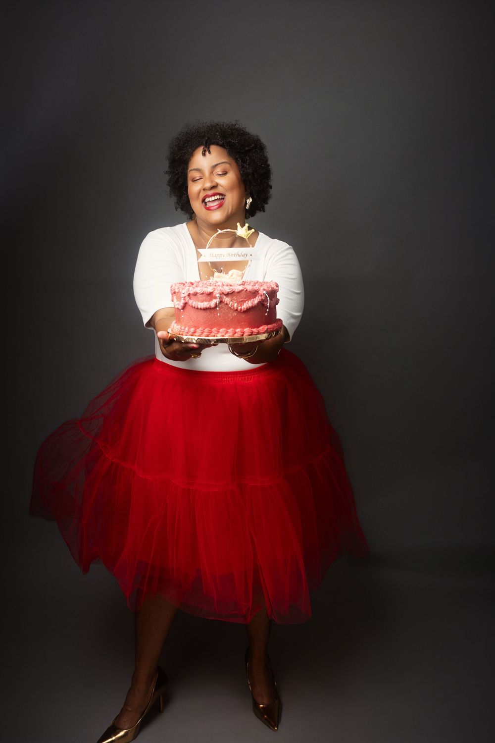 Someone wearing a casual white top and dramatic red tulle skirt holds a pink birthday cake while smiling joyfully.