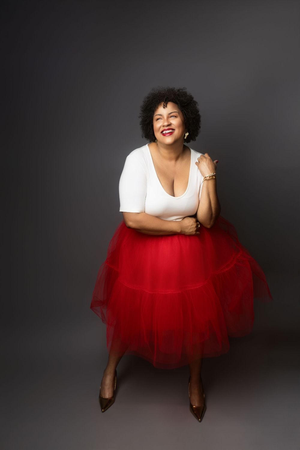 A person wearing a white top and flared red tulle skirt laughs joyfully against a dark gray backdrop.