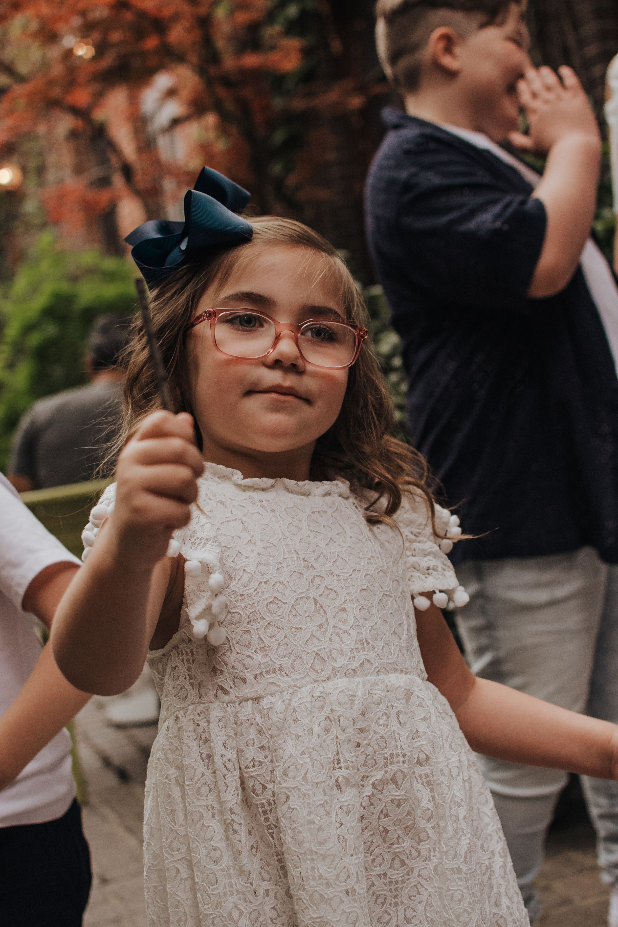 Young child wearing white lace dress and glasses holds up decorative wand at outdoor gathering.
