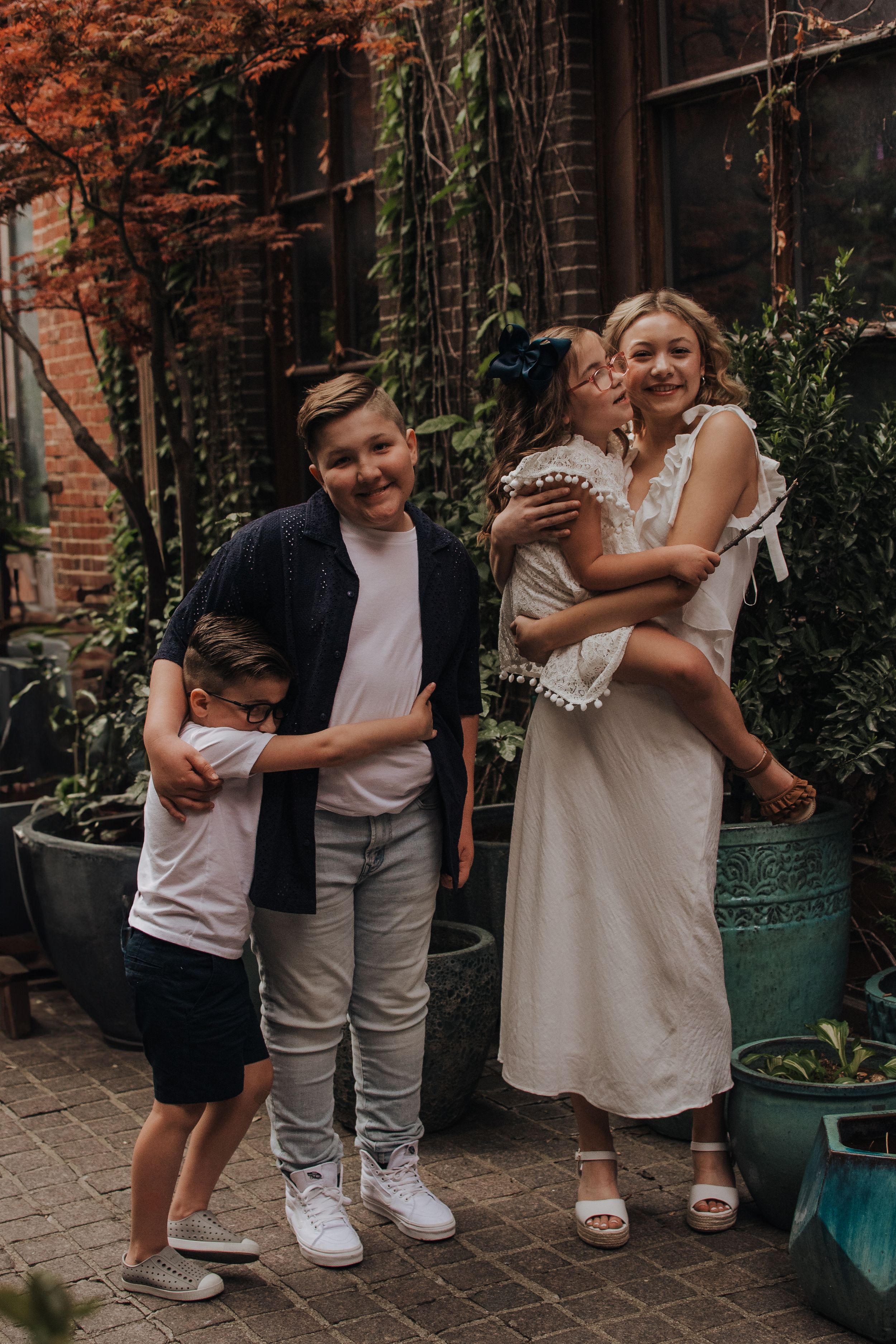 Sweet candid moments captured between siblings sharing hugs and laughter in a garden courtyard setting.