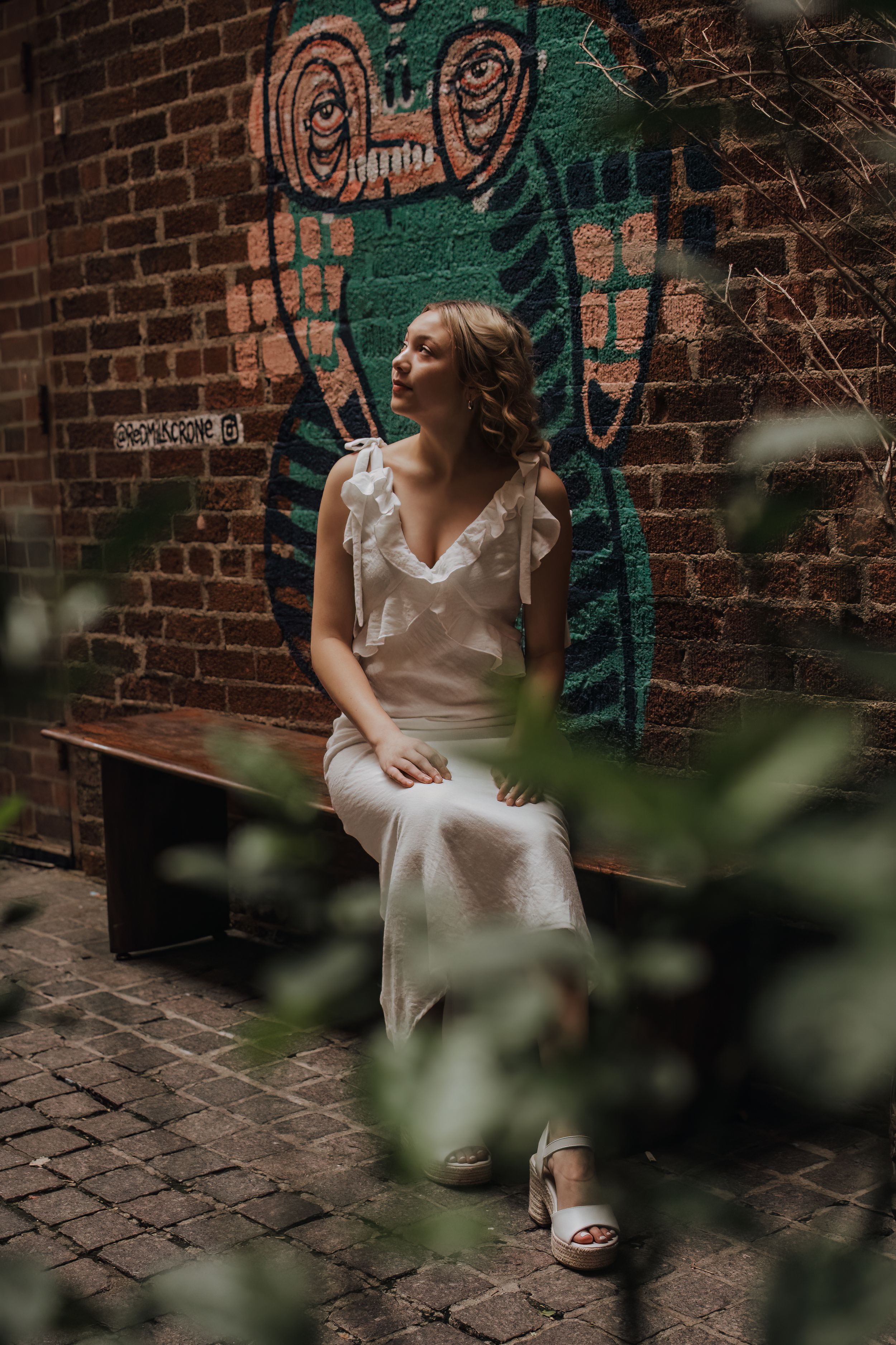 Person in white dress sits thoughtfully against brick wall with green street art backdrop in urban setting.