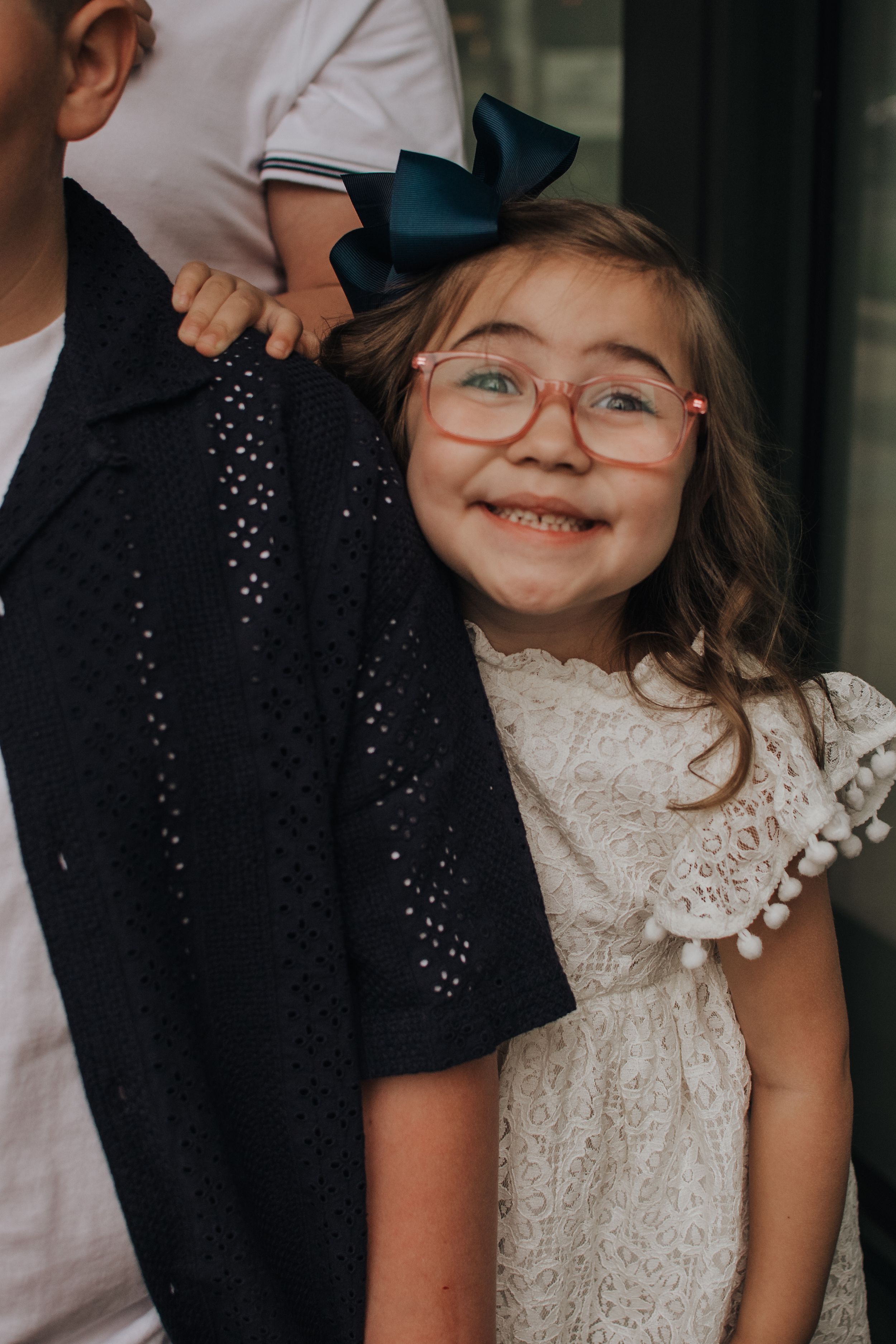 Child in white lace dress and glasses smiles joyfully during outdoor celebration.