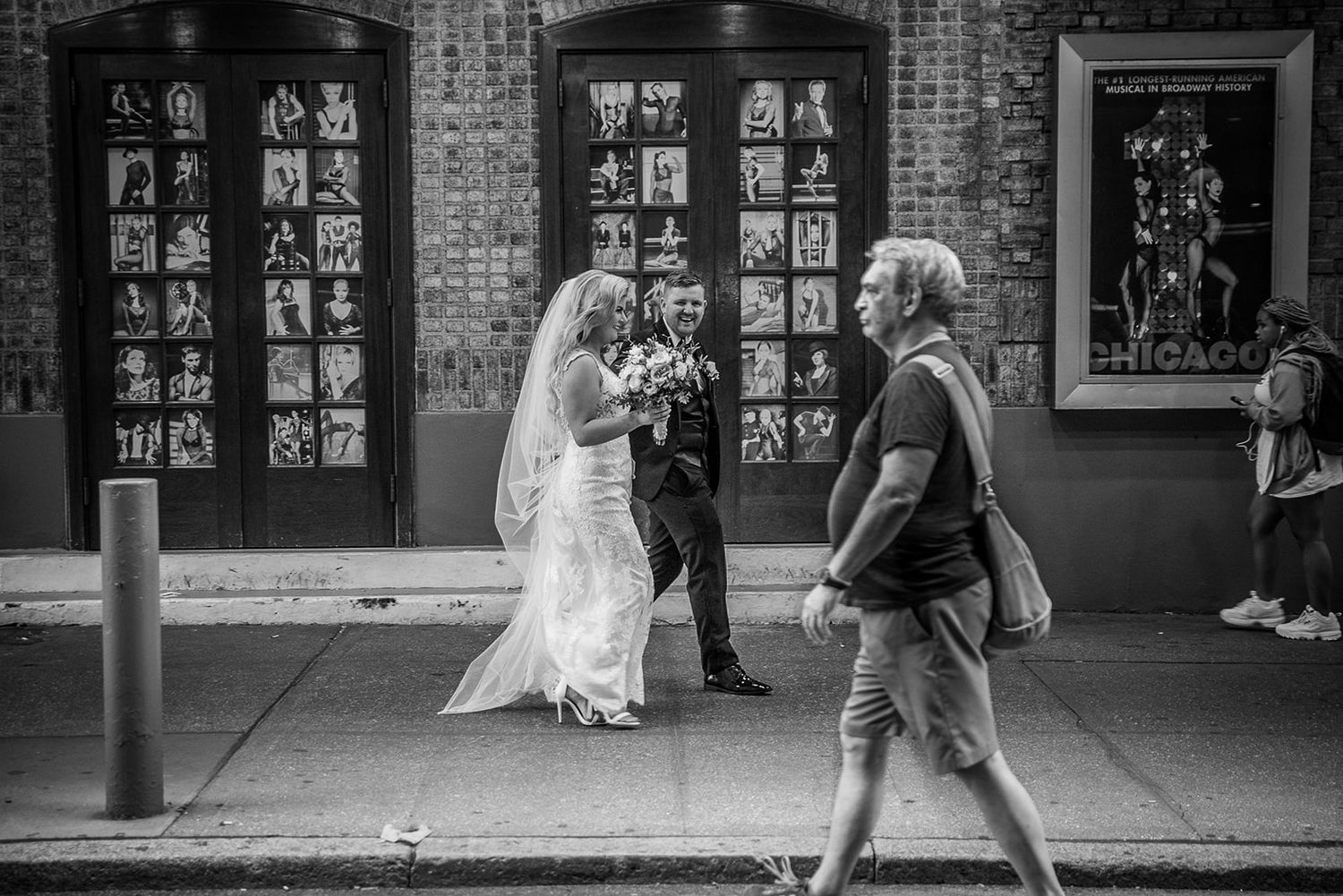 Two people walk past brick storefronts covered with black and white photographs along a city street.