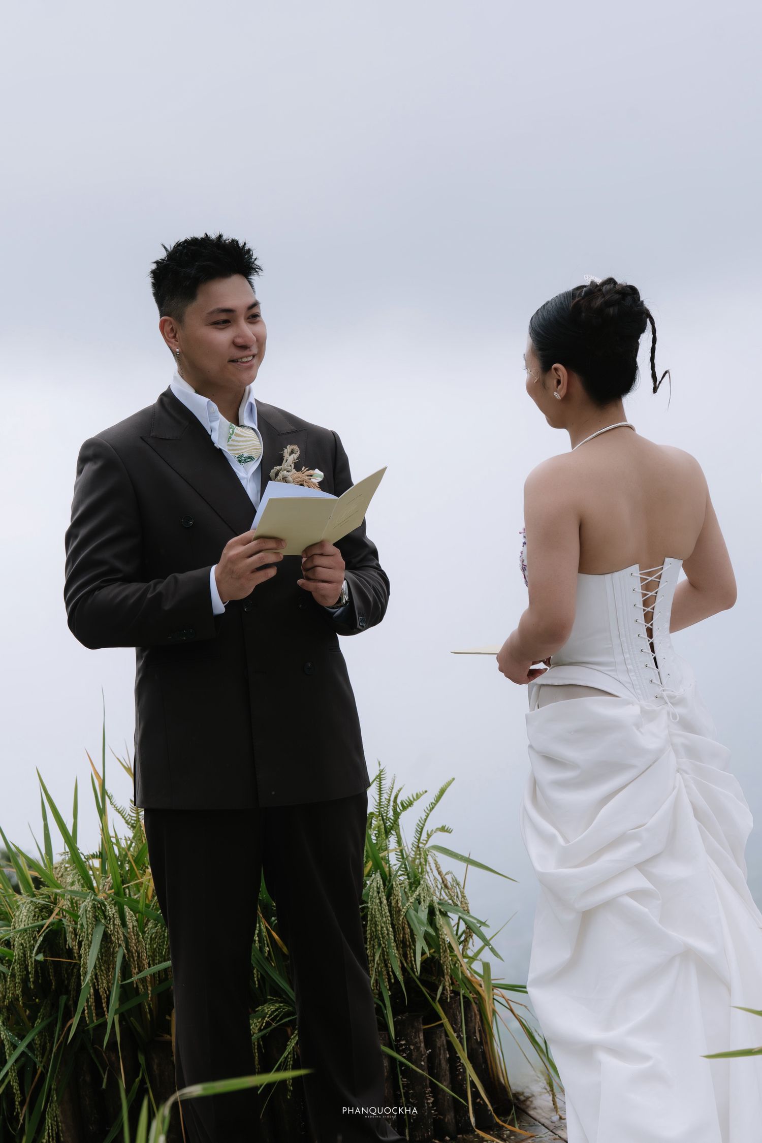 Groom reads wedding vows to bride in white corset dress outdoors in green rice field.