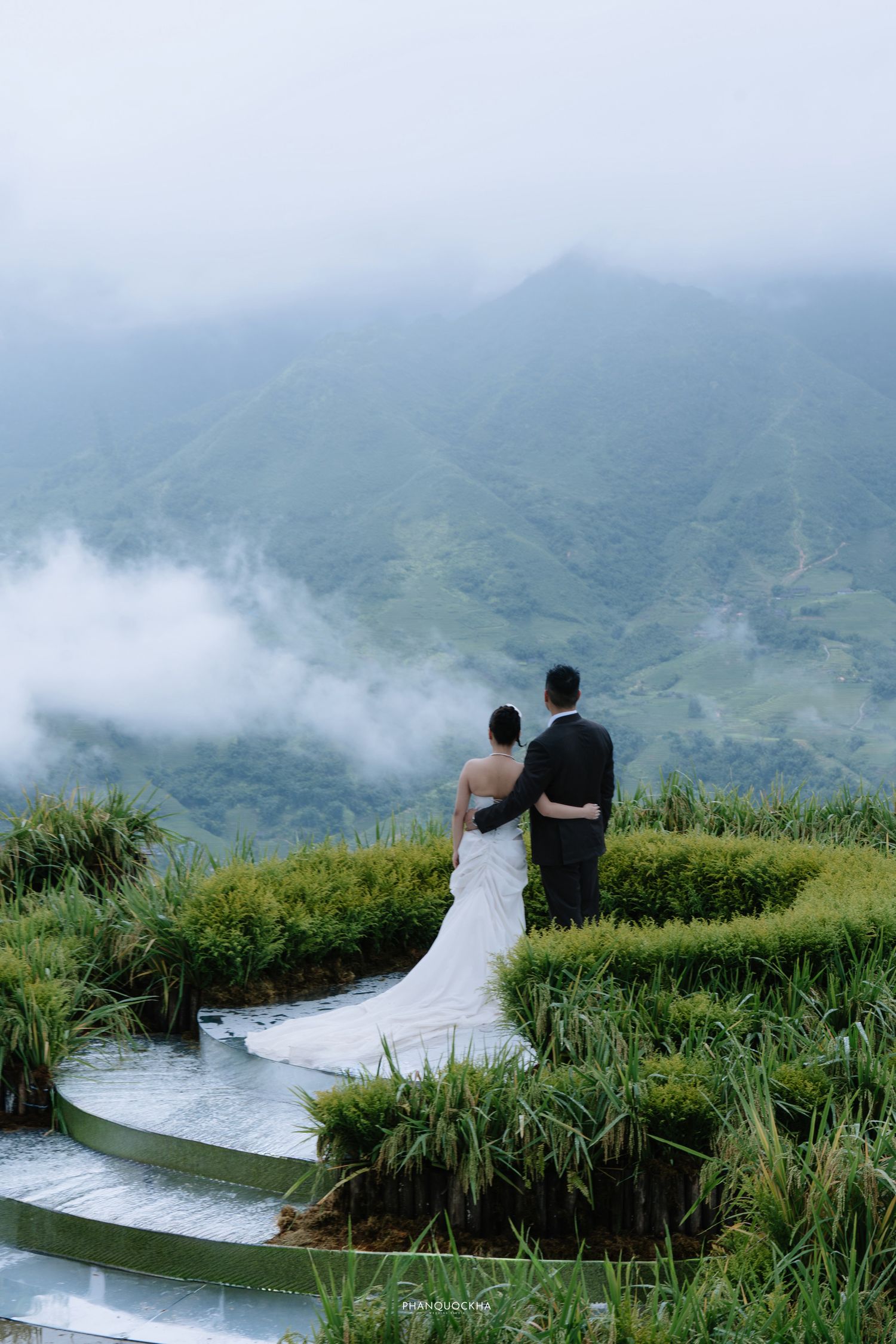 Wedding couple embraces on terraced rice field overlooking lush green misty mountain landscape.