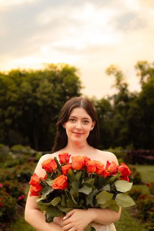 Person holding a beautiful bouquet of fresh coral-colored roses in a garden setting at sunset.