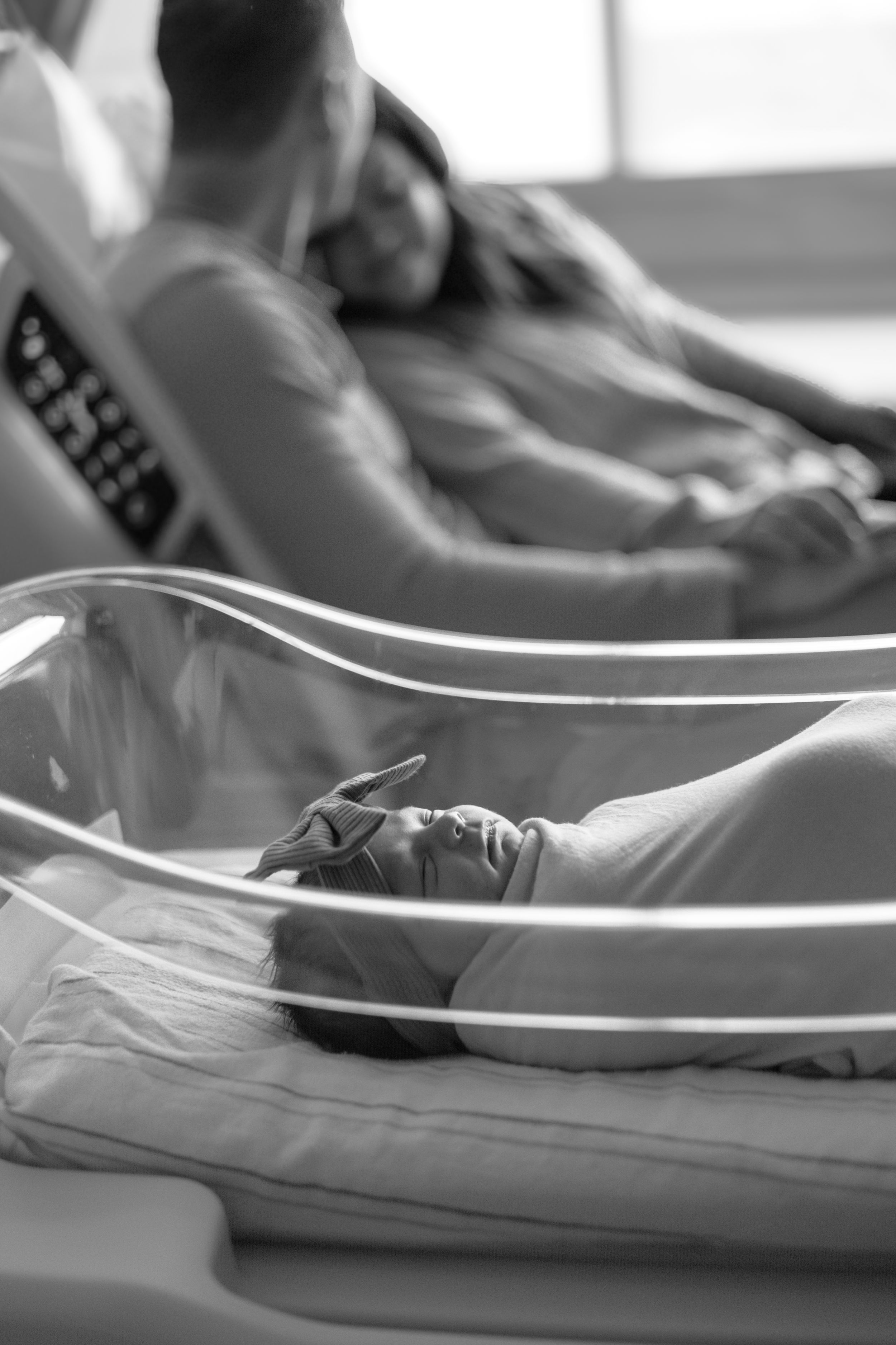 Black and white photo showing a newborn baby sleeping in a transparent hospital bassinet.
