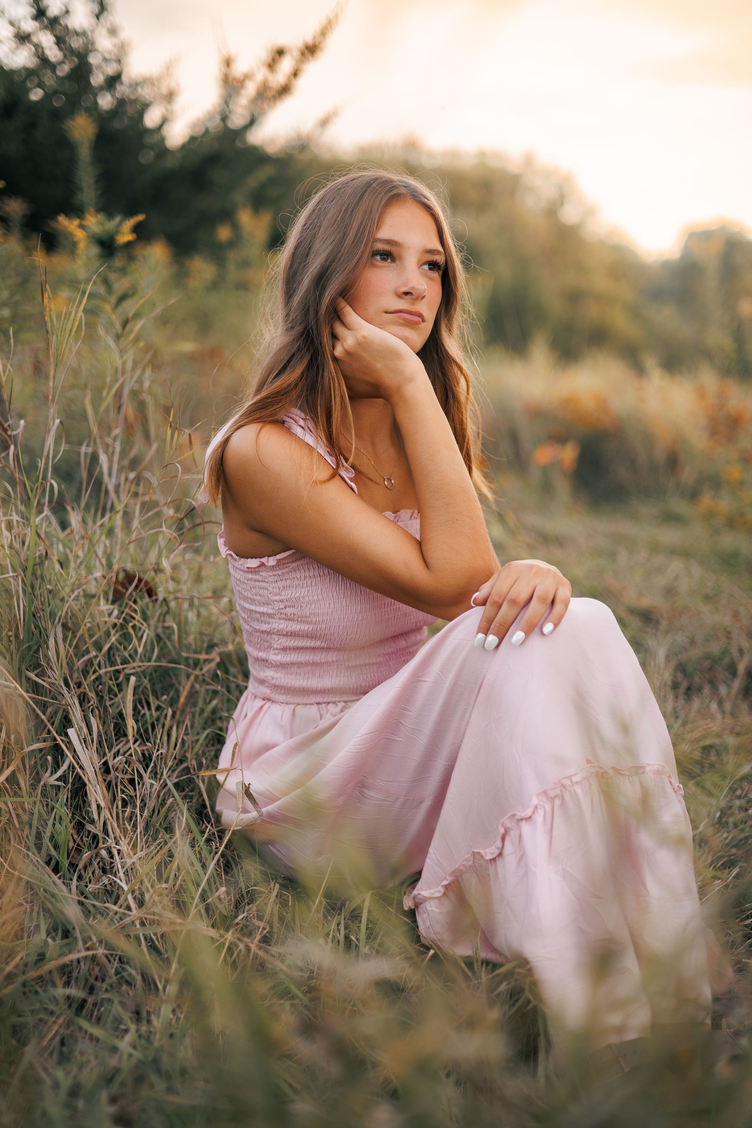 A person in a pink dress sits thoughtfully in a sunlit field of dried grass and wildflowers at golden hour.