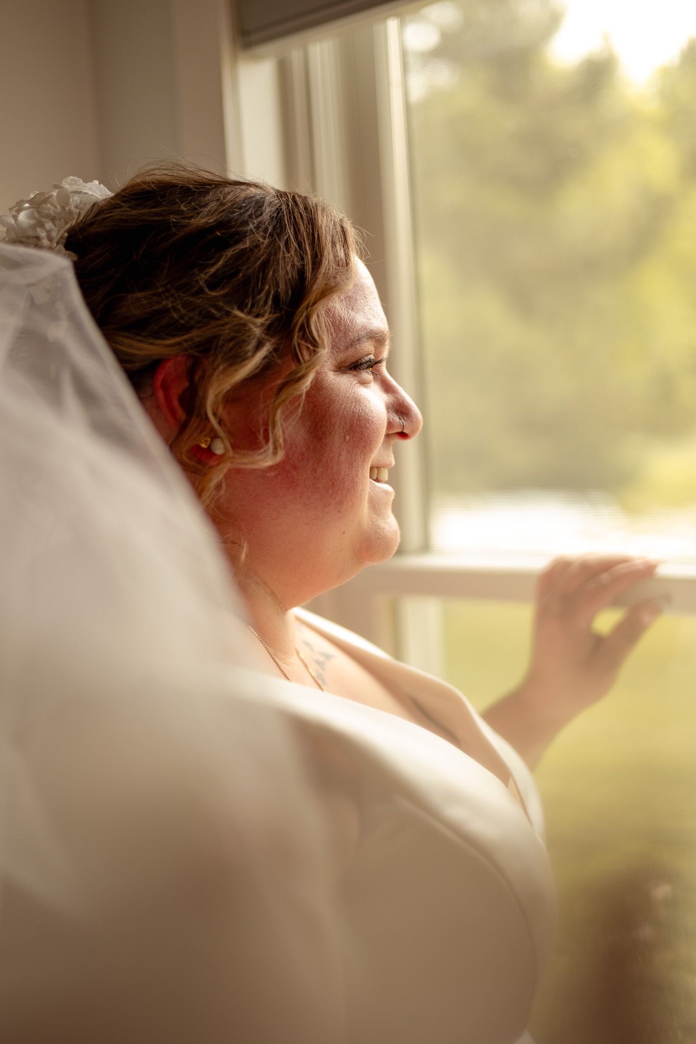 Smiling bride in white gown and veil gazes out a sunlit window on her wedding day.