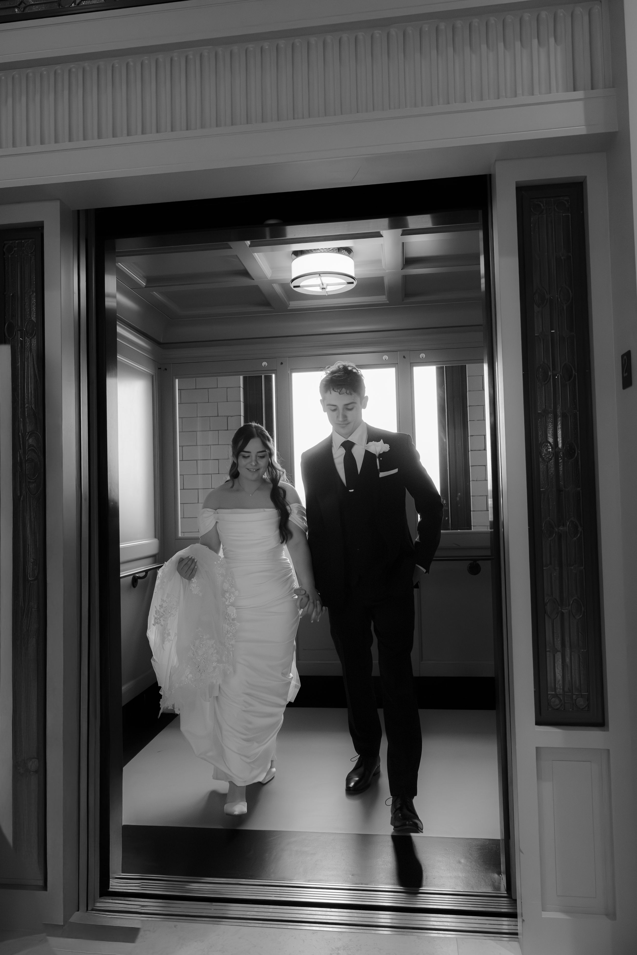 Black and white photo of a bride and groom exiting an elevator, bride in white gown, groom in dark suit.