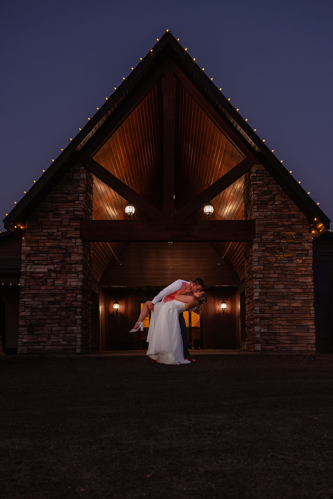 Groom lifting bride outside a rustic stone chapel with warm lights and string lights at dusk.