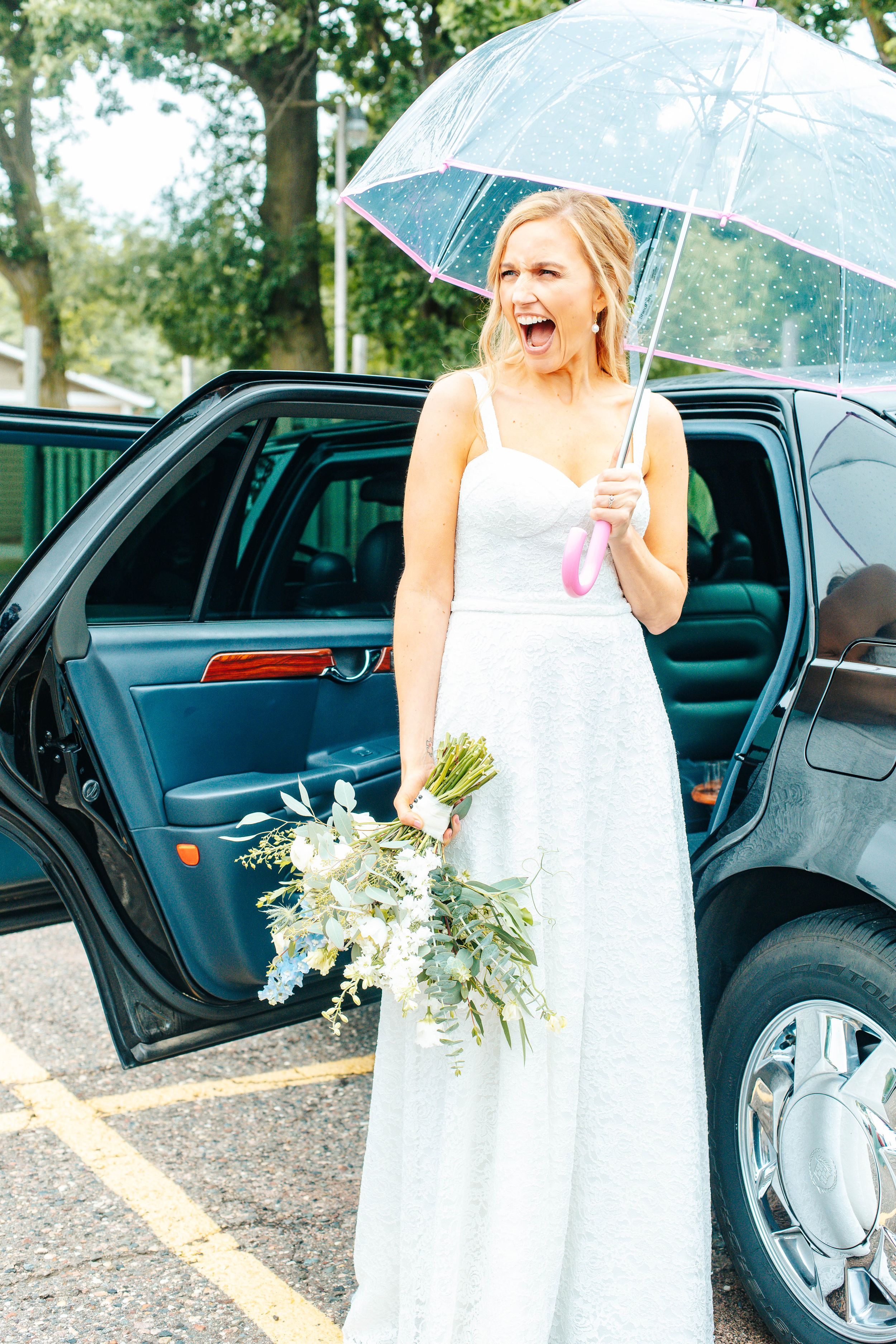 Bride in white gown holds clear umbrella and bouquet, laughing beside black car on rainy wedding day.