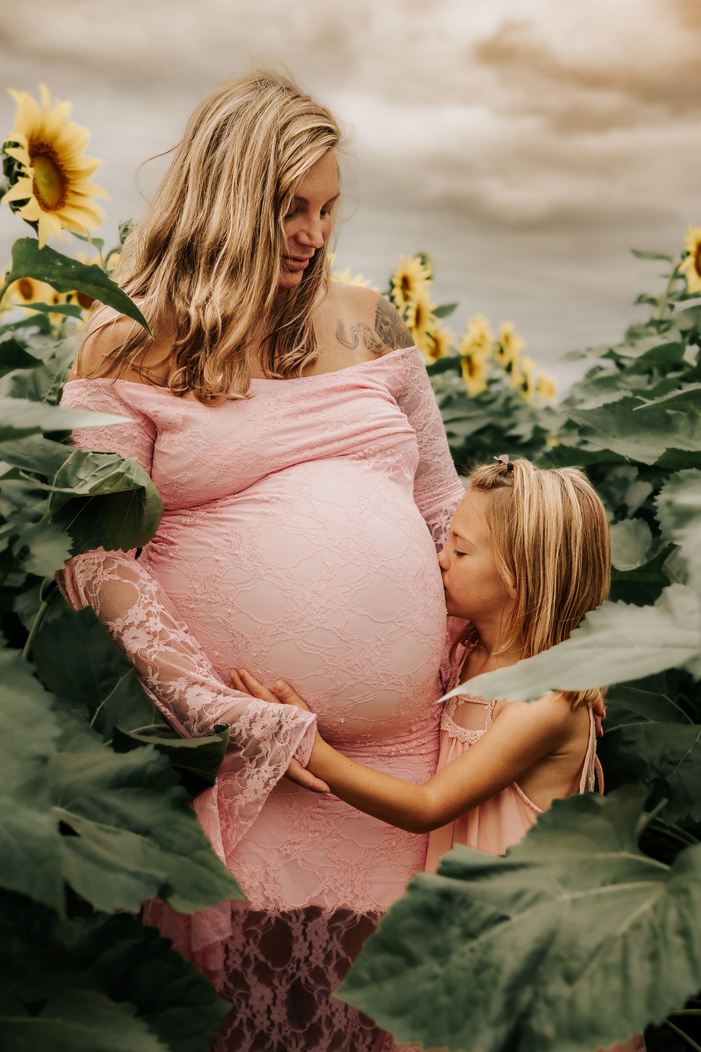 Pregnant woman in pink lace dress with daughter kissing her belly in a sunflower field.