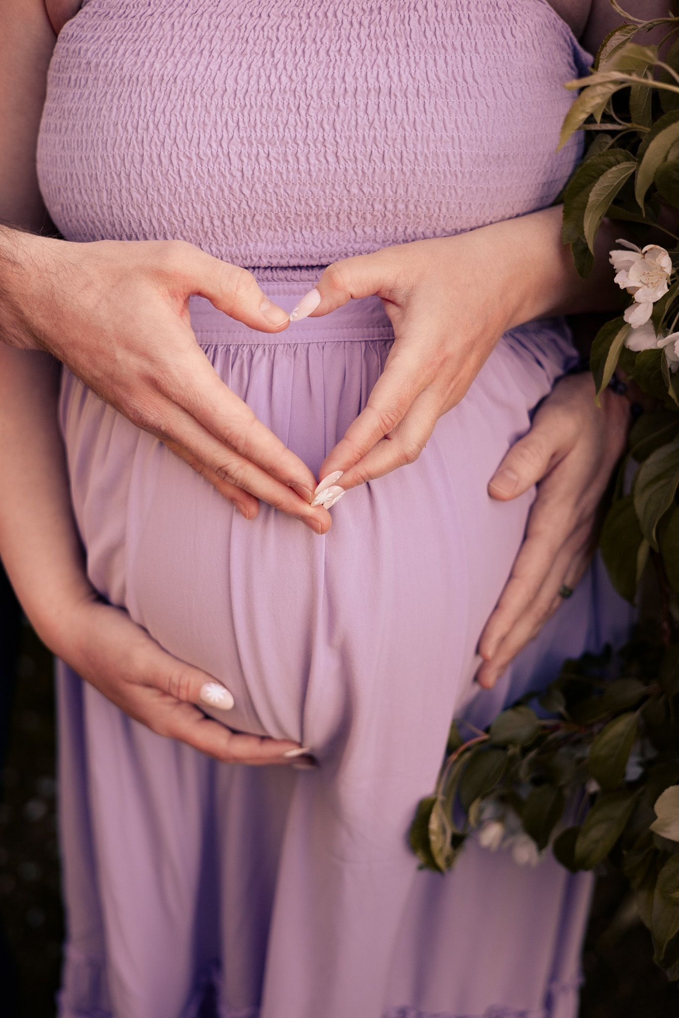 Pregnant woman in lavender dress with partner forming heart shape with hands over baby bump near flowering branches.