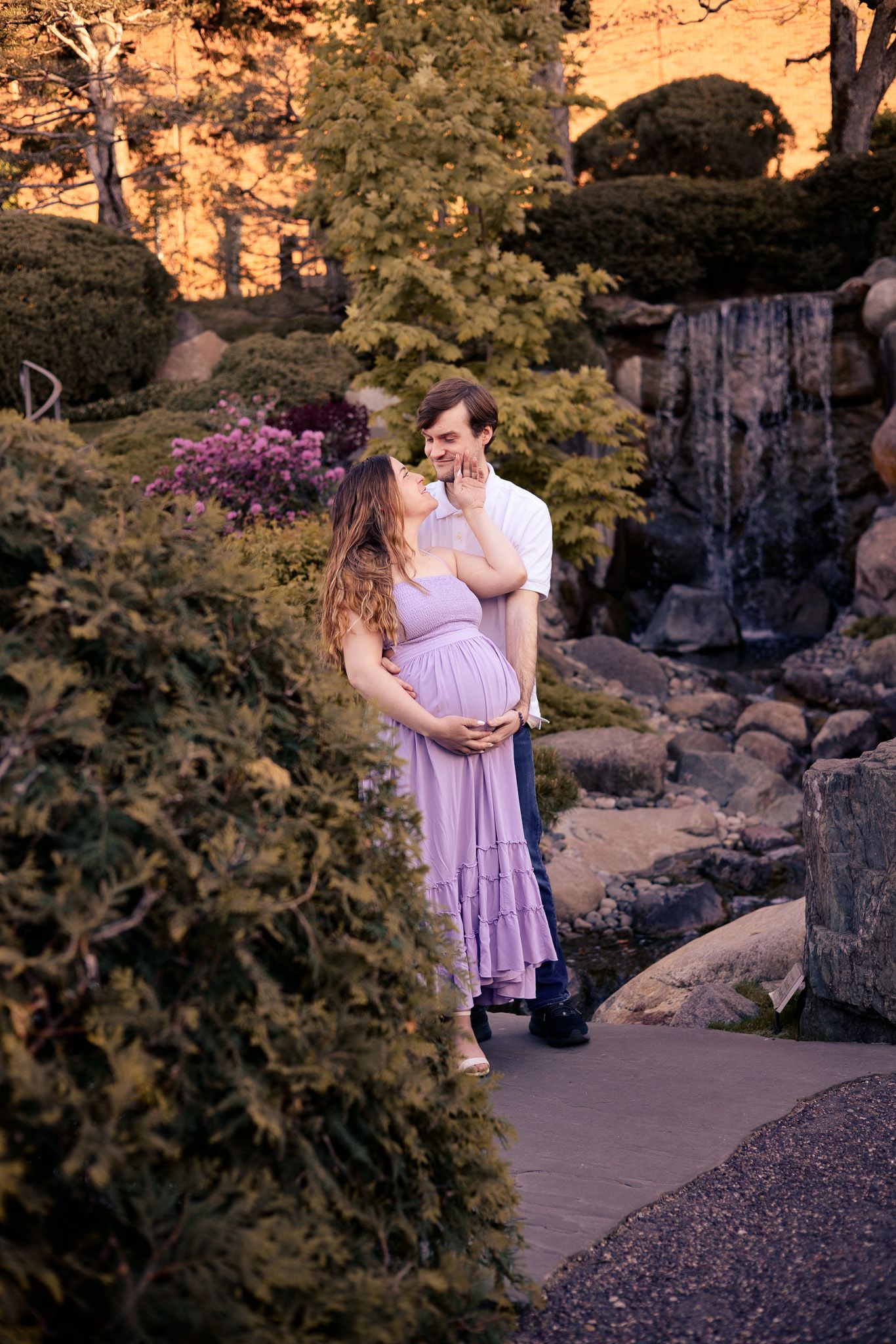 Pregnant couple embracing in a lush garden with a waterfall, woman in lavender dress touching man's face at golden hour.