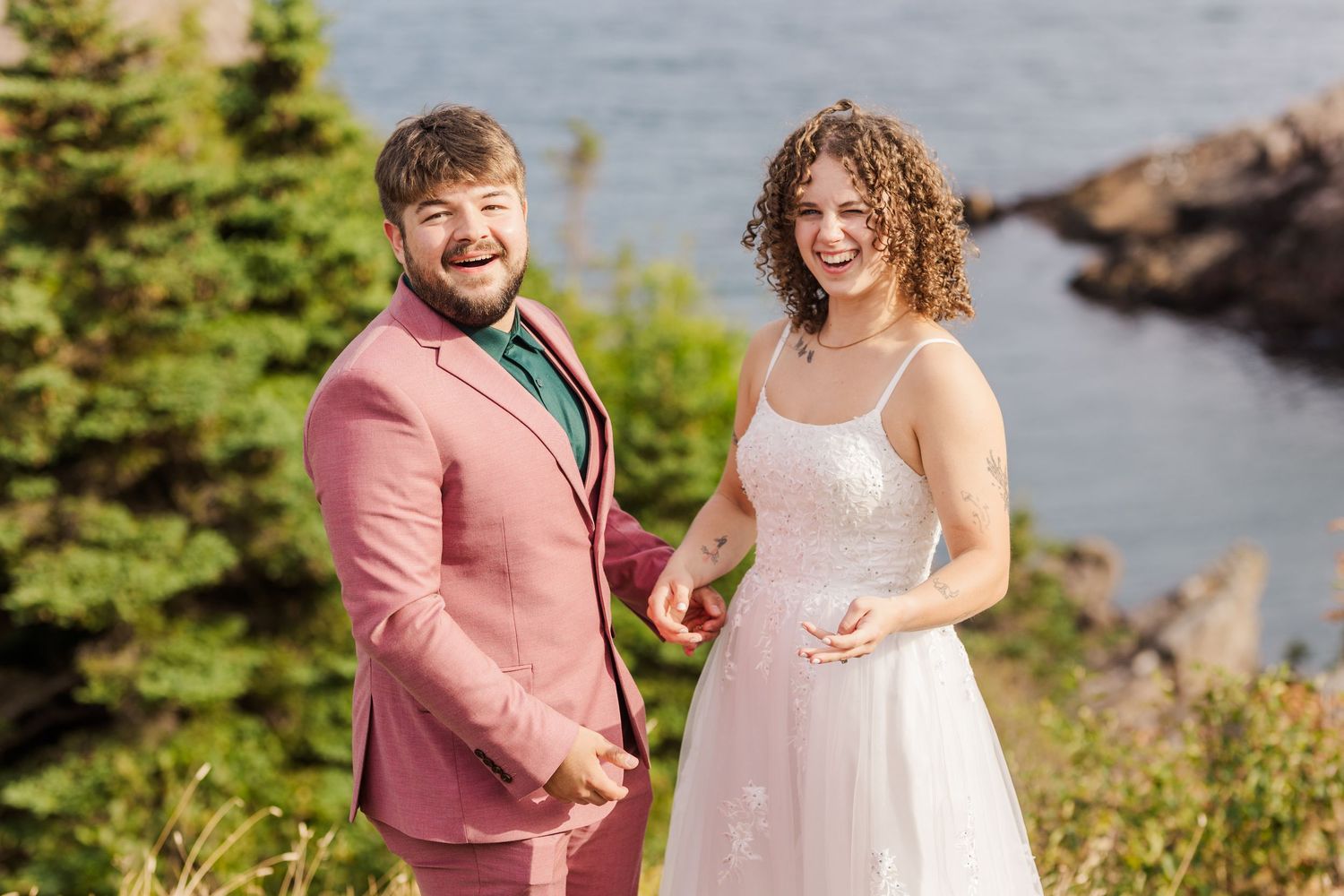 A couple poses happily together outdoors by the water with evergreen trees in a scenic coastal location.