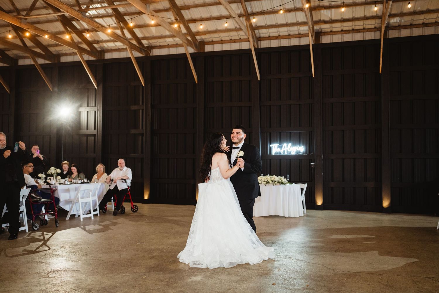 A bride and groom share their first dance in a rustic wooden barn with dramatic lighting and guests watching from the sides.