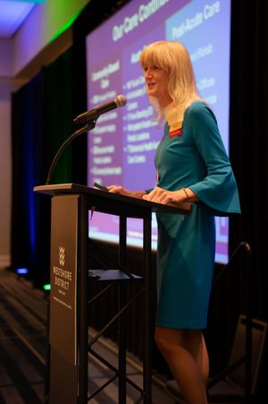 Speaker presenting at a podium during a professional conference with projection screen in background.