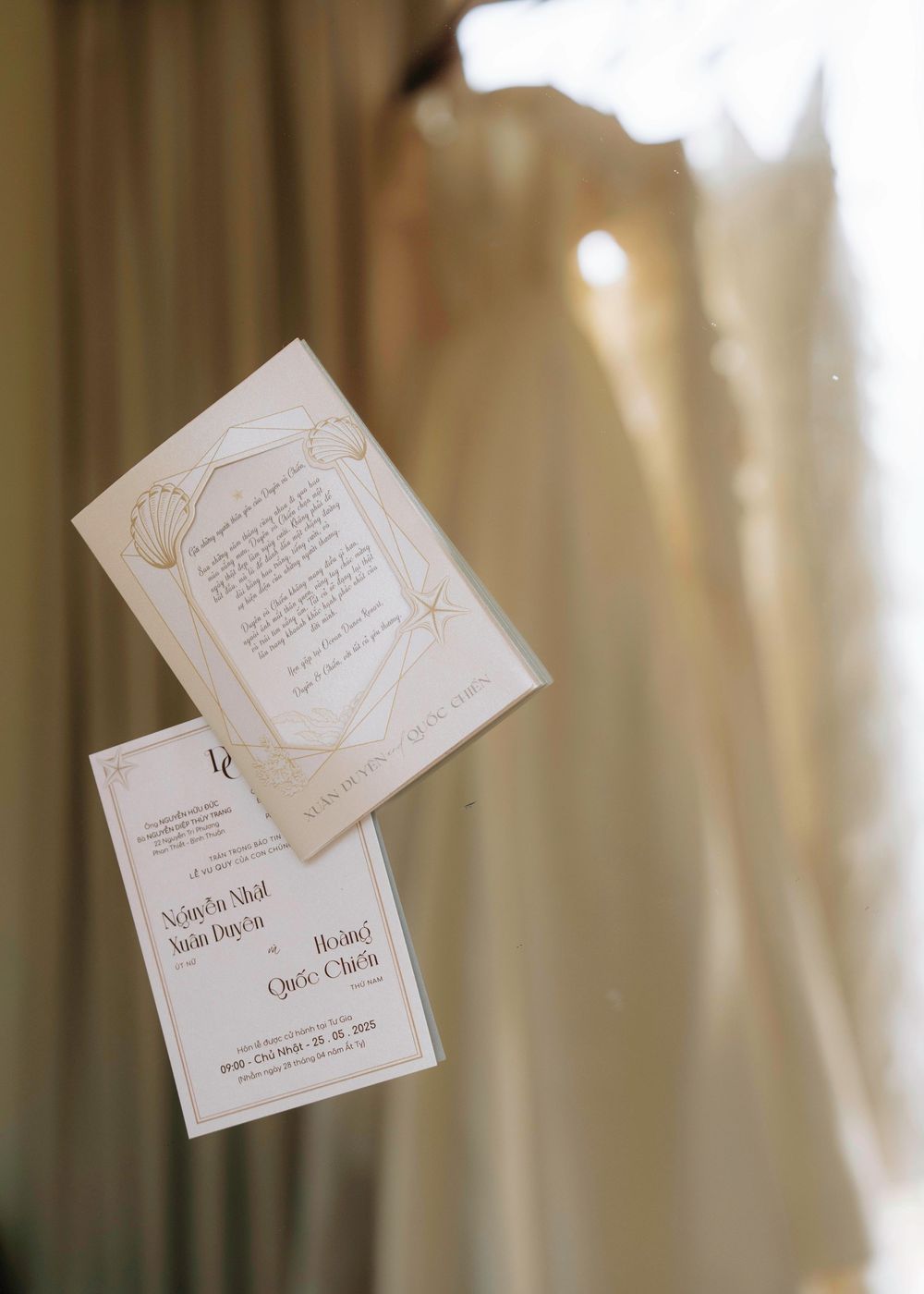 White wedding invitation cards floating in front of a hanging bridal gown near sheer curtains.